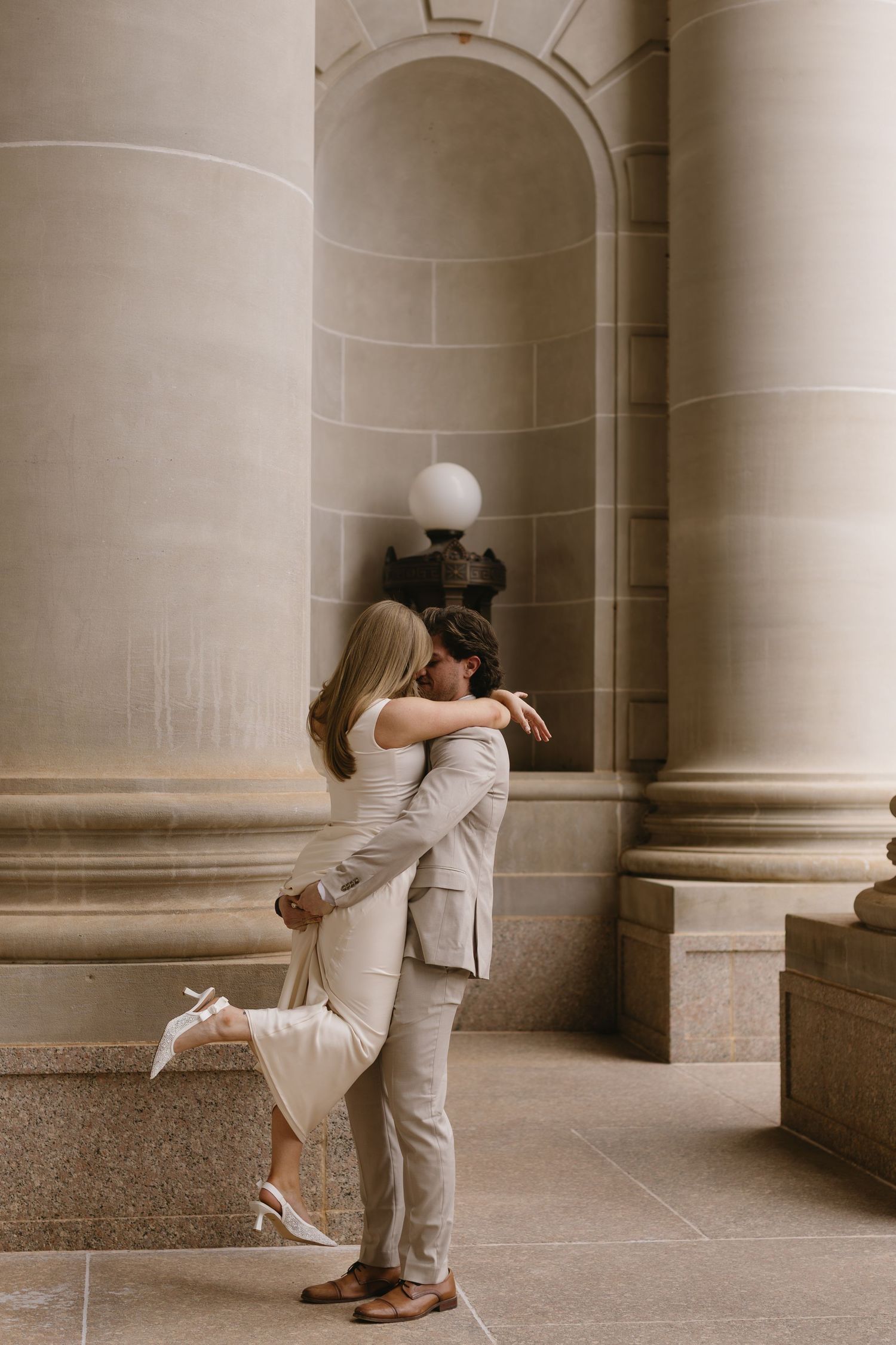 A couple in formal wear embraces in a colonnade with classic architectural details.
