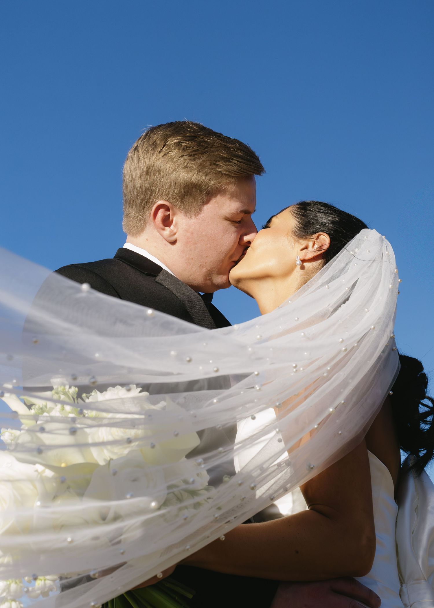 Romantic wedding kiss with flowing veil against bright blue sky.