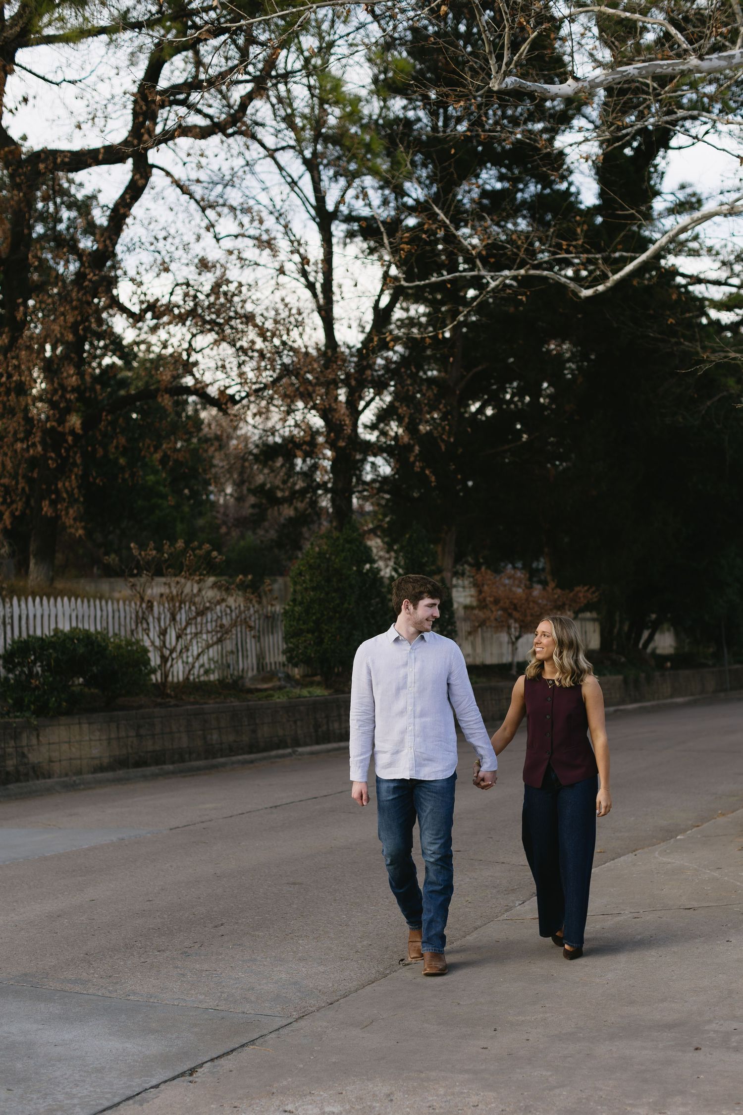 A couple walks hand in hand along a paved pathway surrounded by trees at sunset.