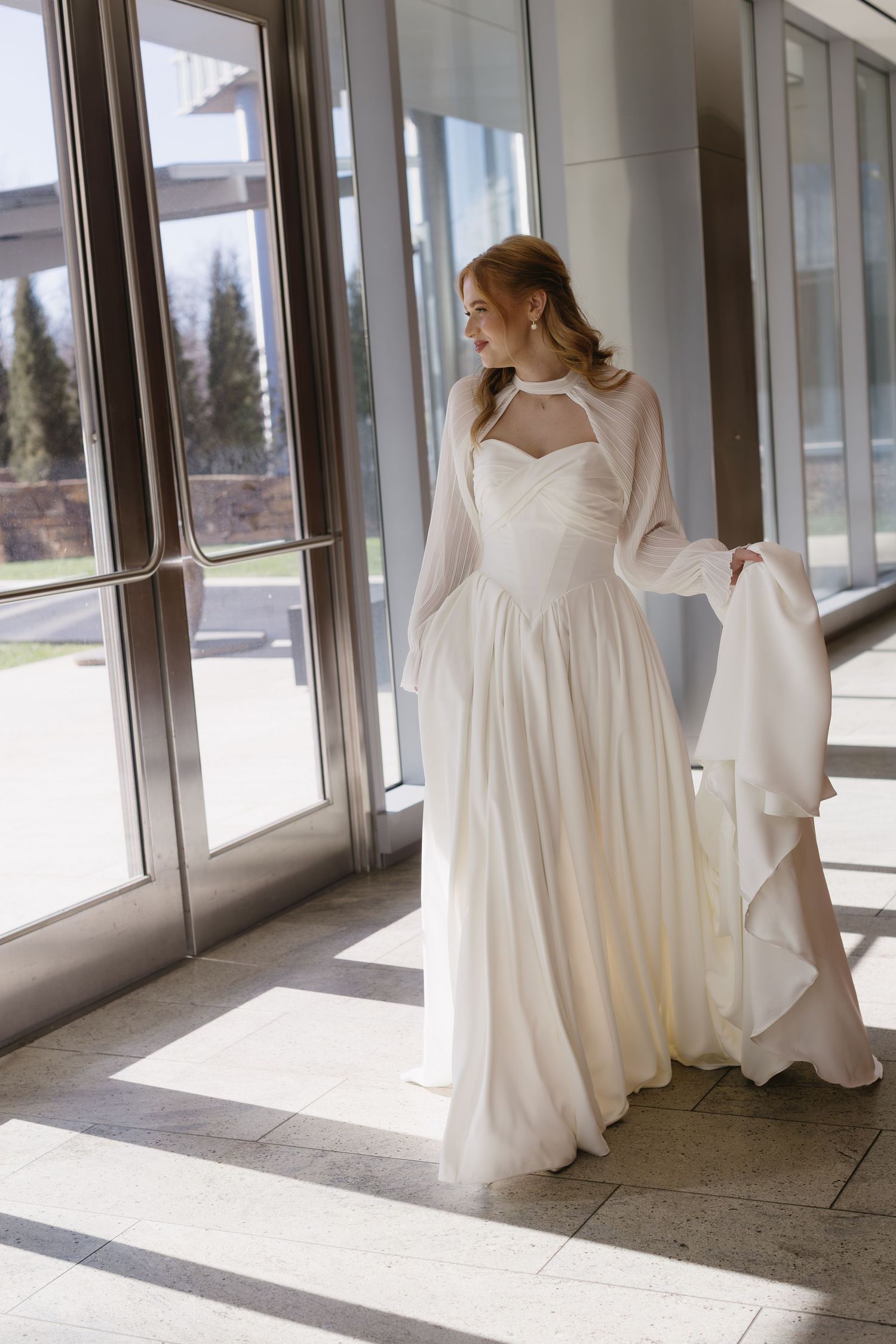 A bride in a flowing white dress twirls gracefully in a sunlit hallway with large windows.