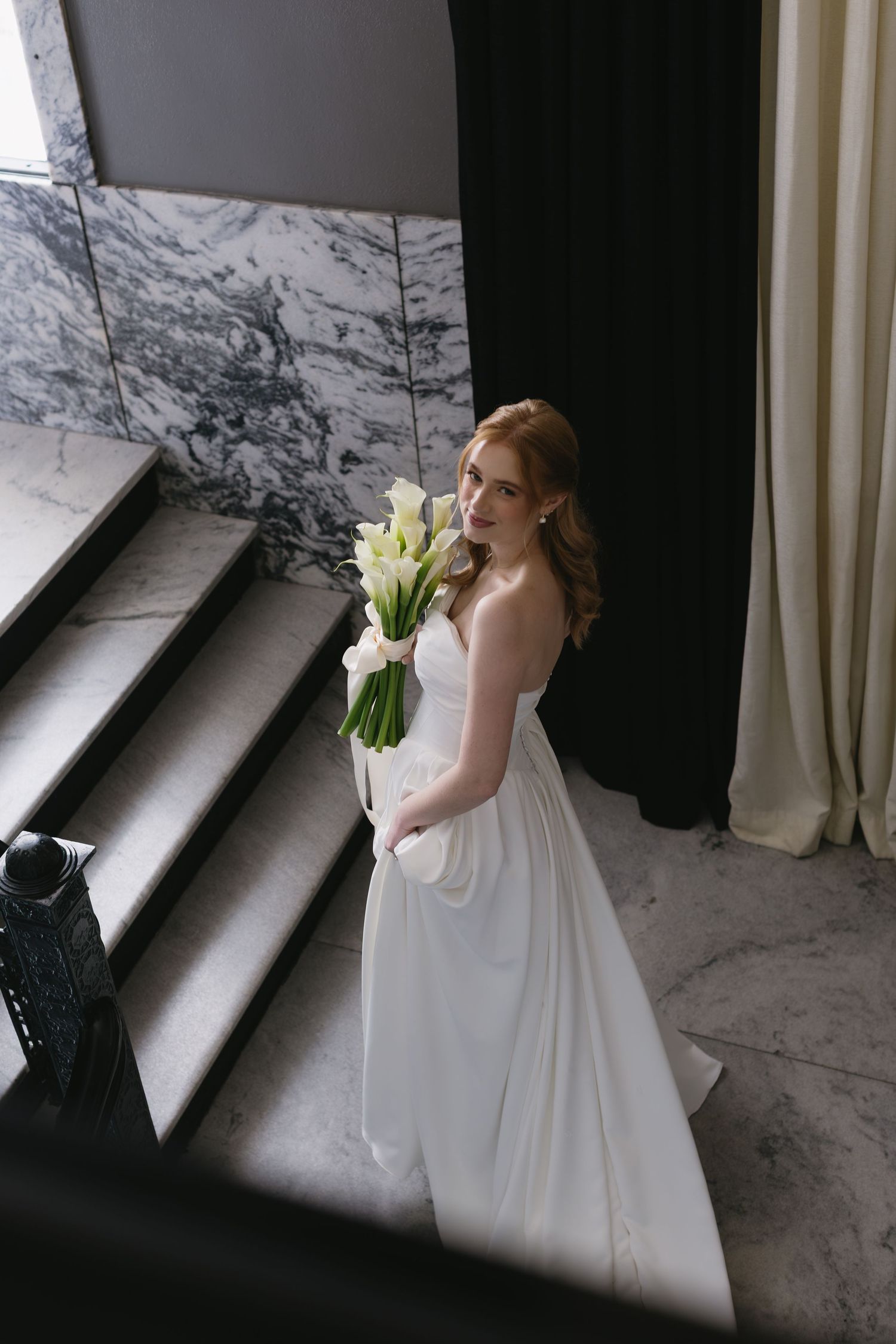 Bride holding white tulips poses by window in flowing wedding gown with dramatic lighting.