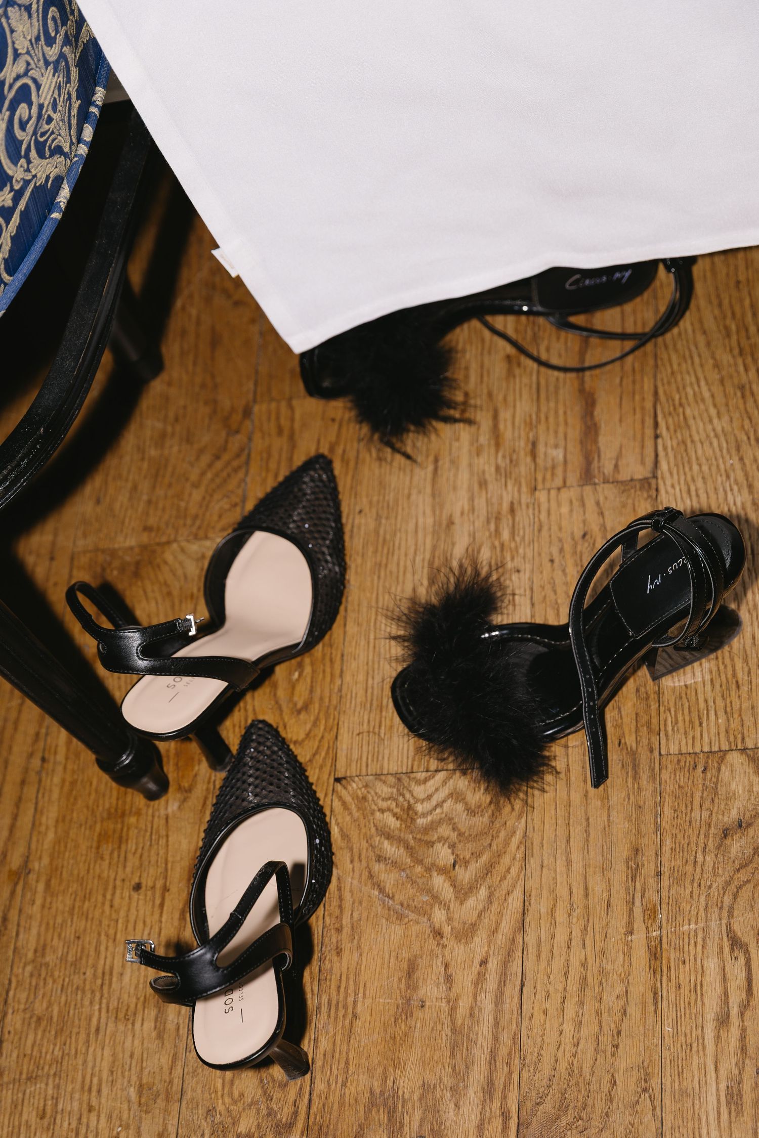 Black feathered high heel shoes on a wooden floor next to white fabric.