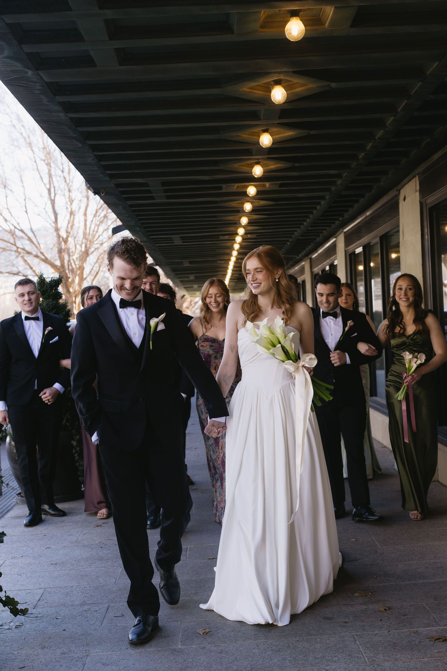 Wedding party walking and laughing together under covered walkway with overhead lighting.