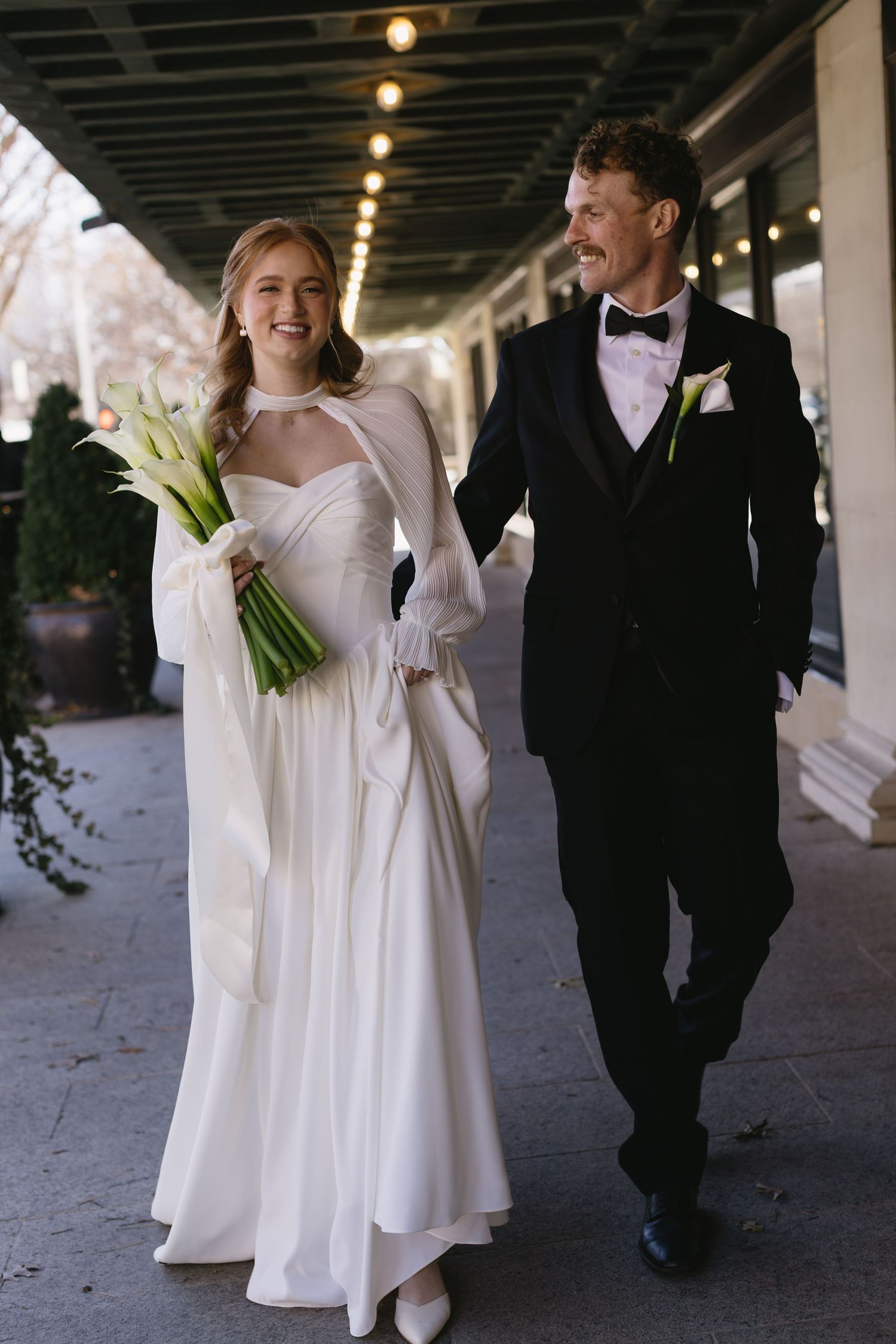 Bride in elegant white dress and groom in tuxedo walking together under covered walkway.
