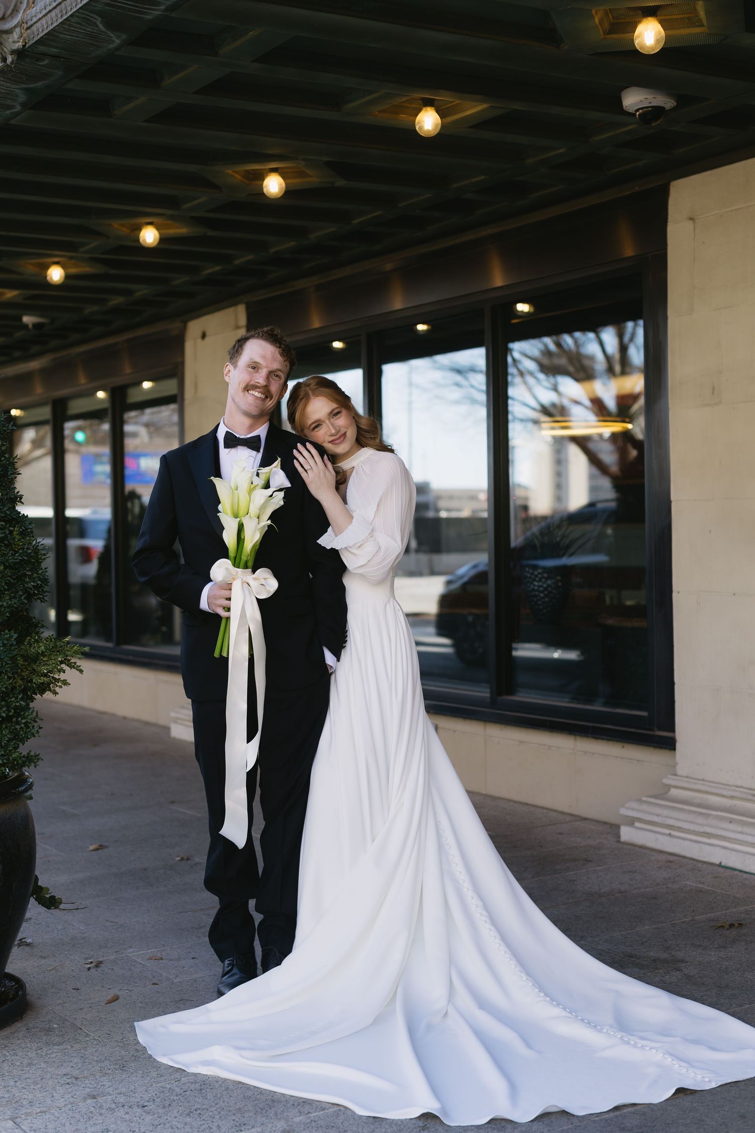 Wedding couple poses outdoors under covered walkway with dramatic train of white dress.