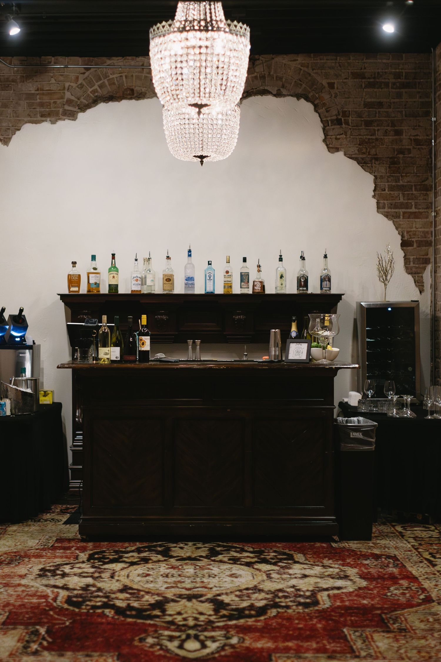 Elegant bar setup with crystal chandelier and liquor display against white and exposed brick wall.