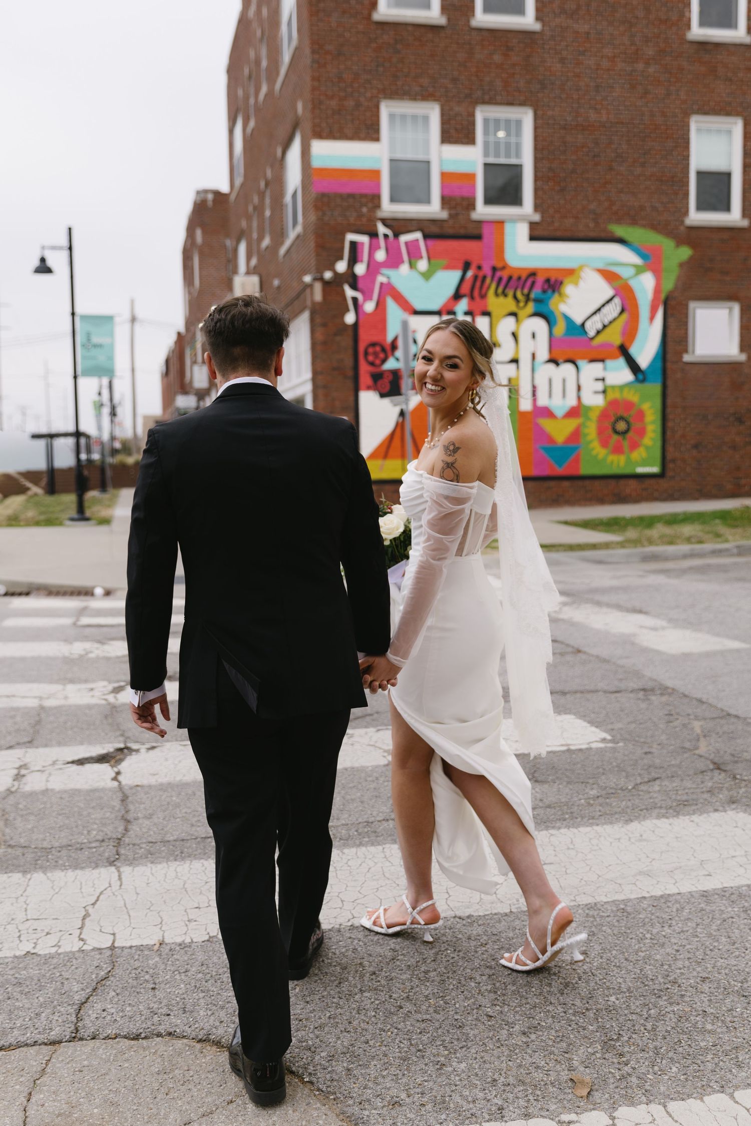 Couple walking across street with colorful mural in background on wedding day.