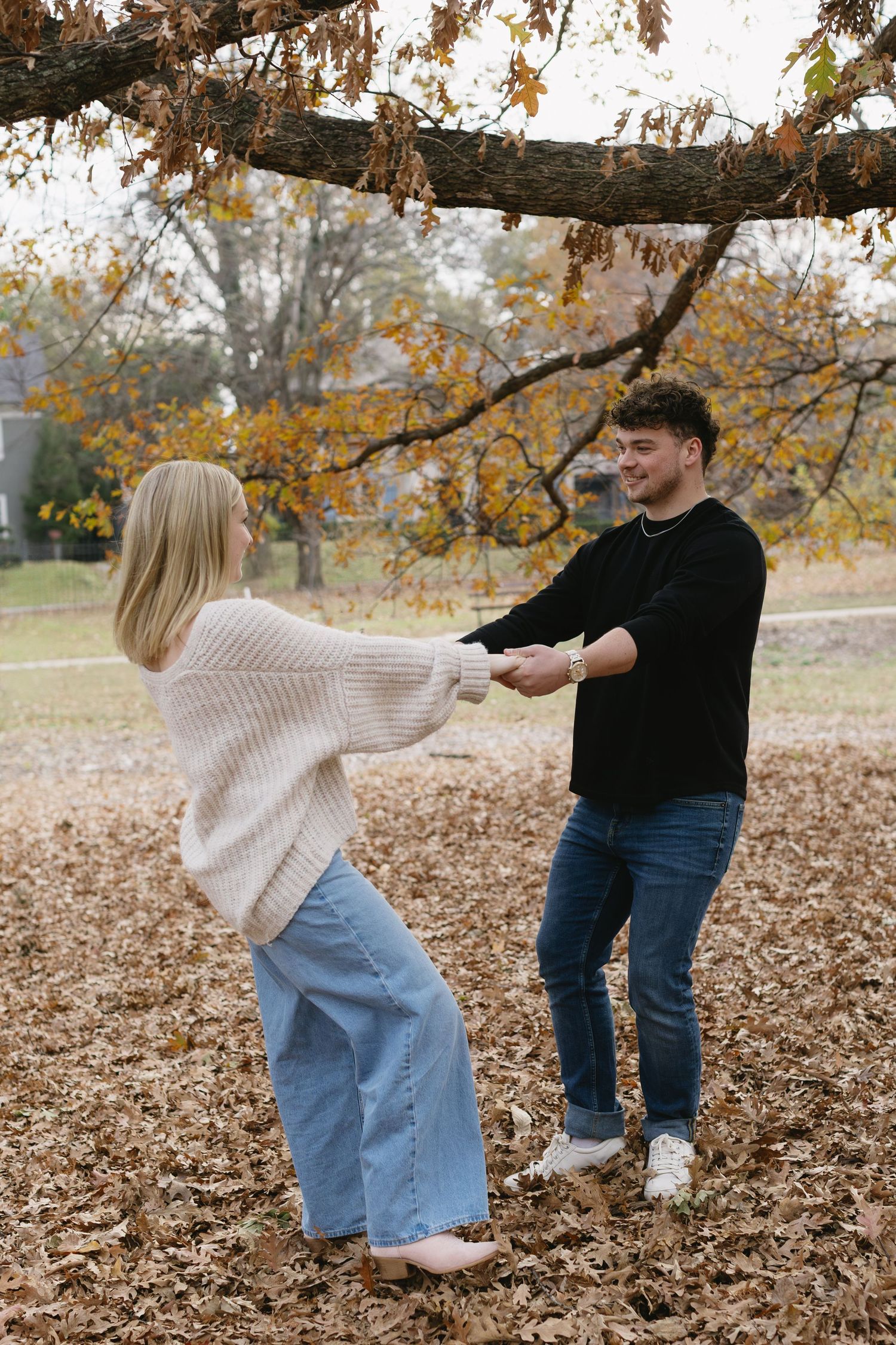 A couple playfully dances together on fallen autumn leaves beneath a tree with golden foliage.