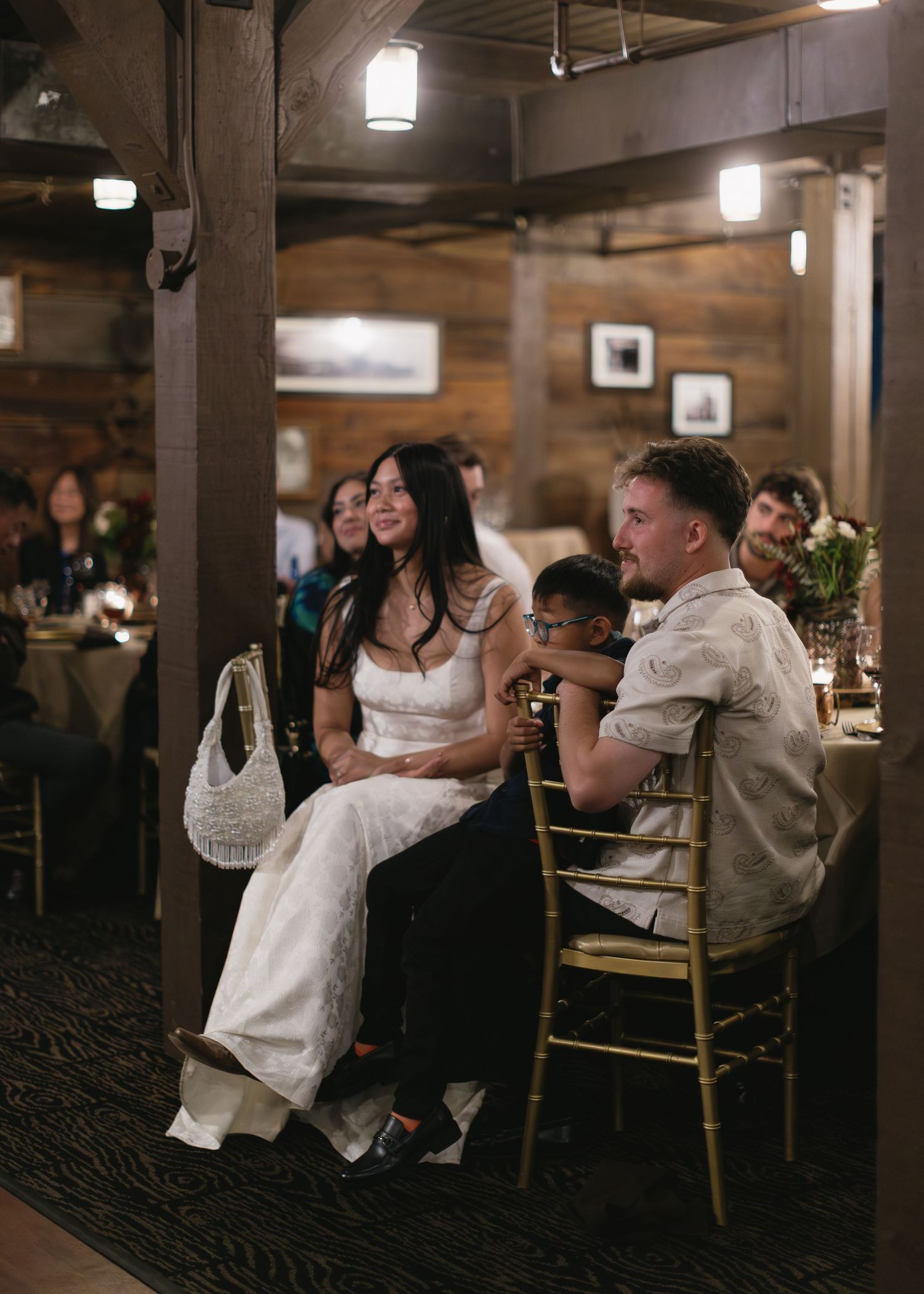 Wedding guests seated at long tables in a rustic wooden venue with pendant lighting.