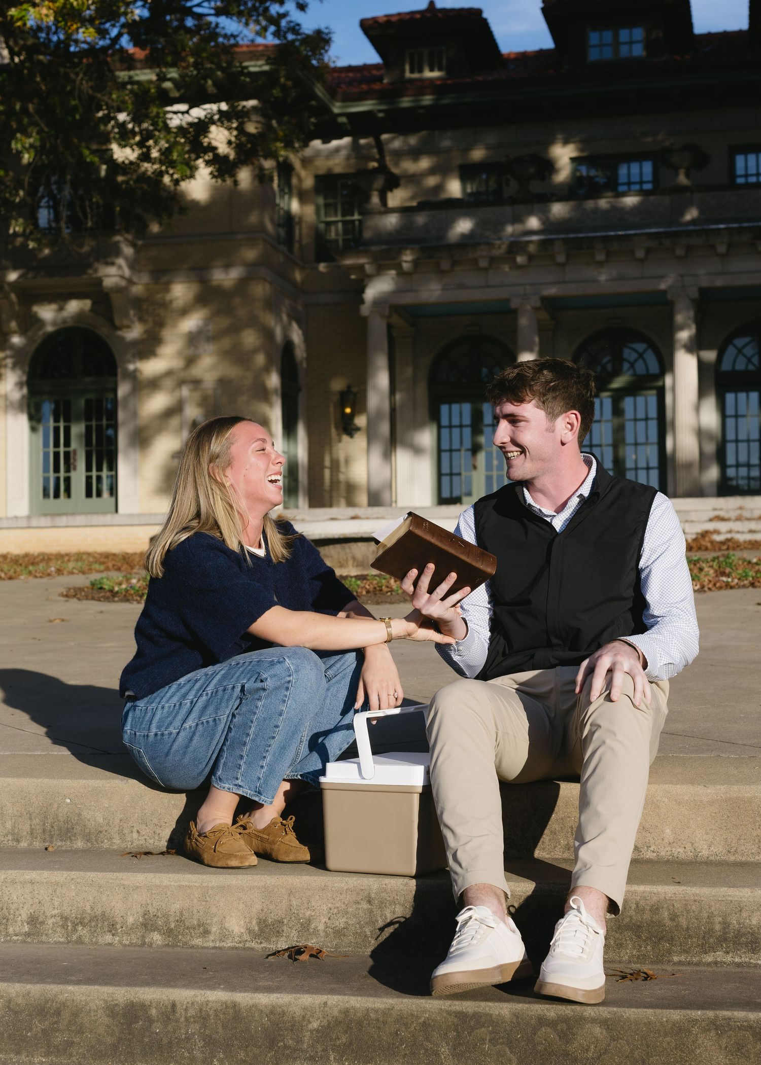 A couple sitting on steps sharing a laugh while looking at a book together.
