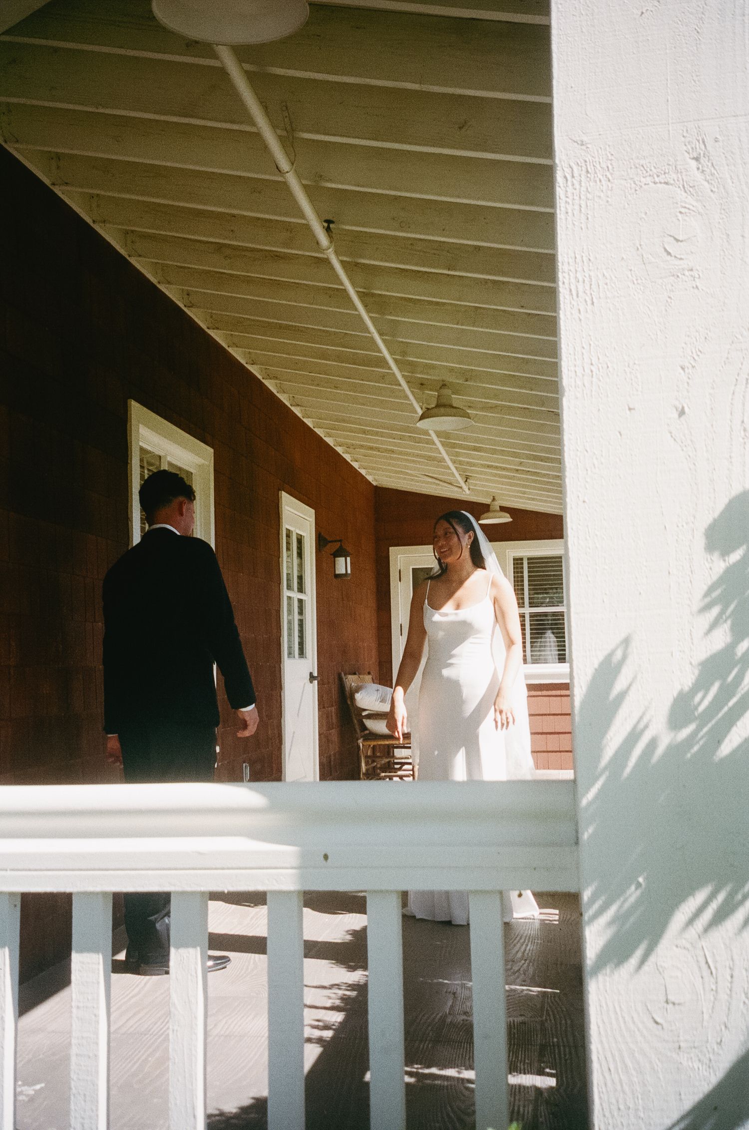A covered porch with white railings and columns casts shadows on a sunny afternoon.