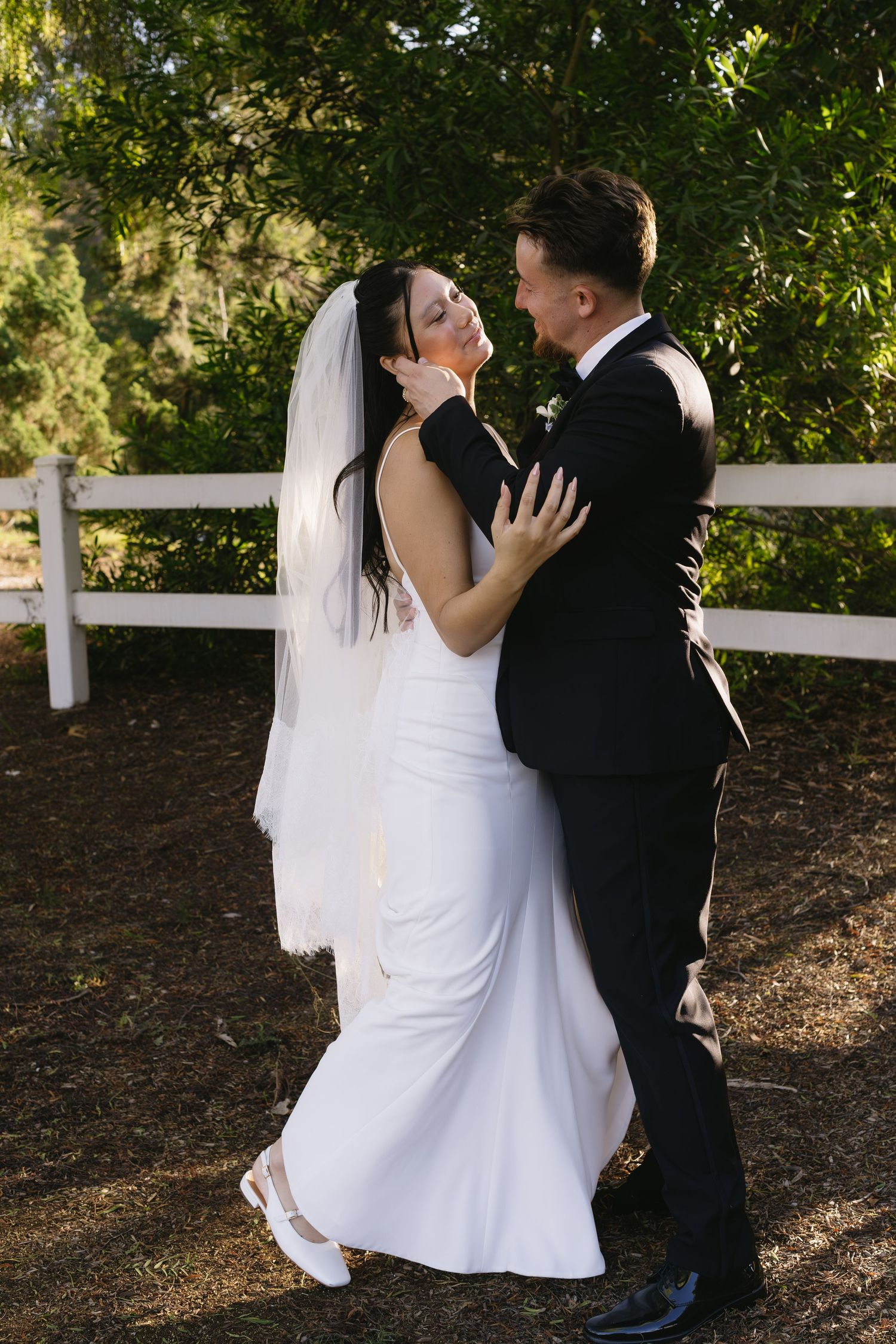 Couple shares intimate moment against white fence during outdoor wedding photoshoot.