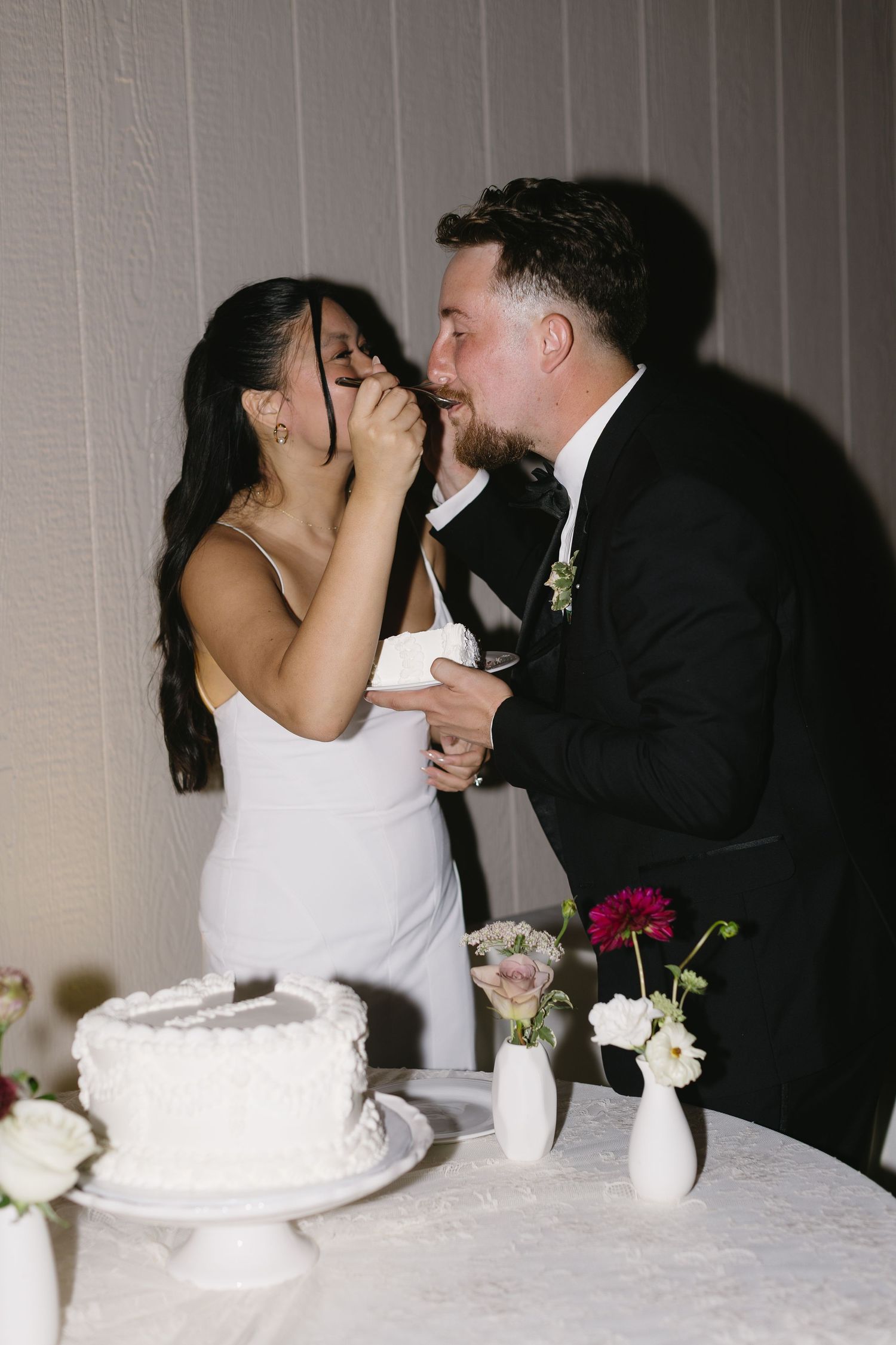 Wedding couple shares their first bite of cake at the reception table decorated with white flowers.