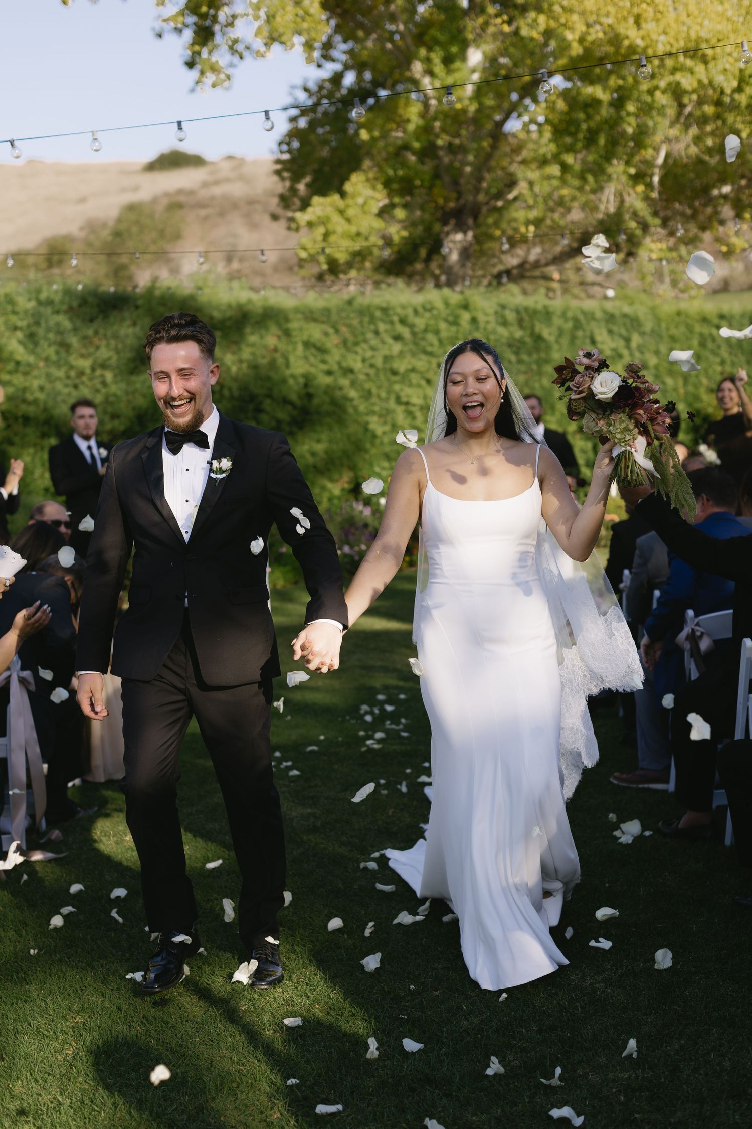 A wedding couple walks through flower petals being tossed by guests during an outdoor ceremony.