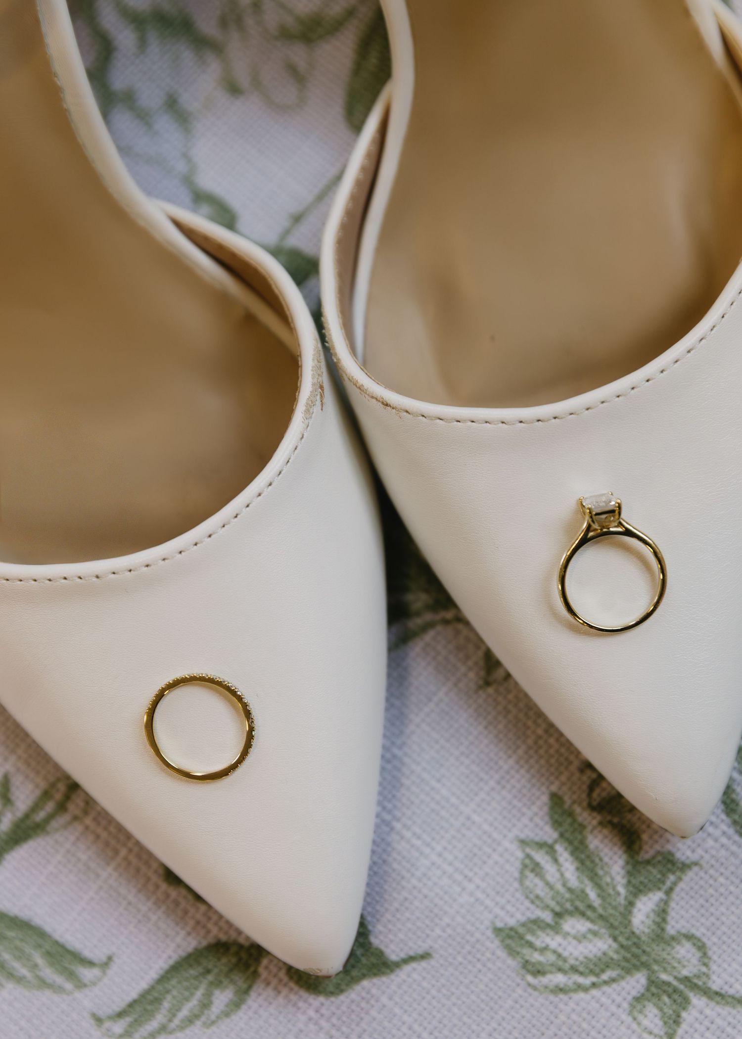 Wedding rings rest elegantly on a pair of cream-colored pointed toe bridal shoes with floral pattern background.