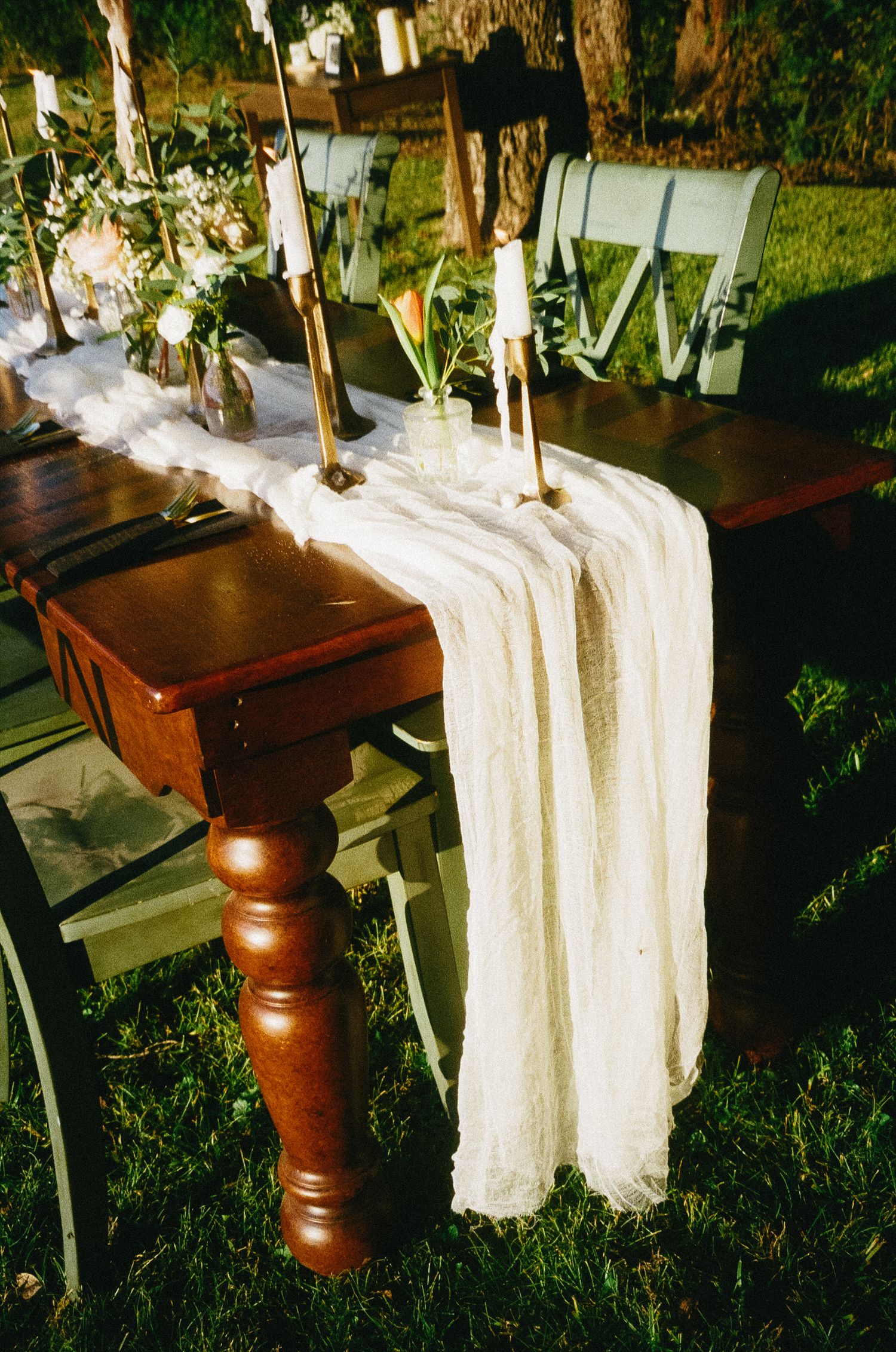A dark wooden table with a white lace runner and candlesticks displayed in an outdoor garden setting.