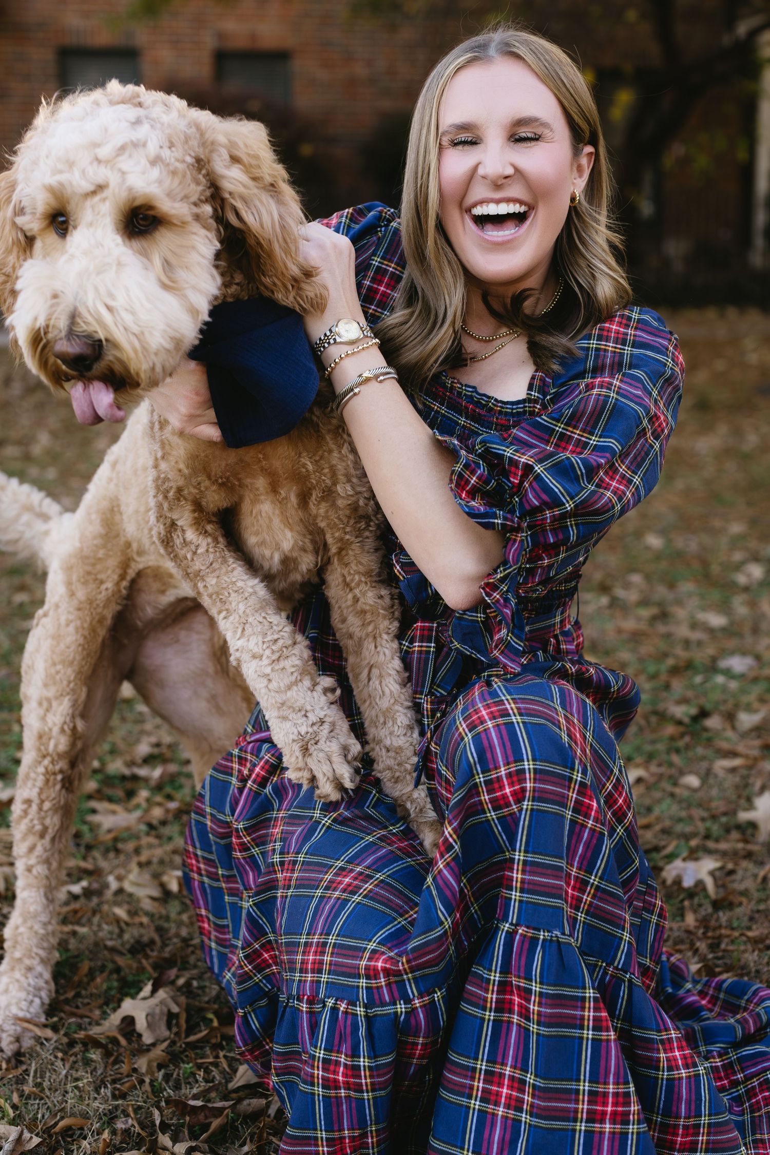 A woman in a plaid dress laughs while hugging her curly-haired Goldendoodle dog outdoors.