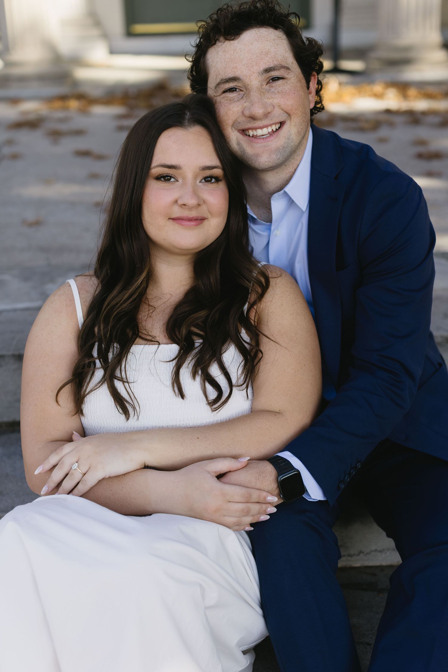 Casual portrait series of couple in white dress and navy suit sitting together outdoors.