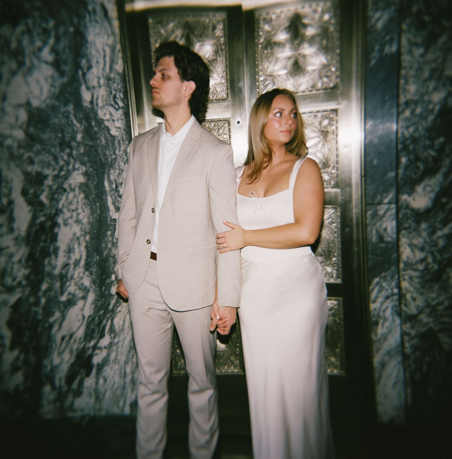 A couple in white and beige formal wear poses against a textured dark wall with ornate details.