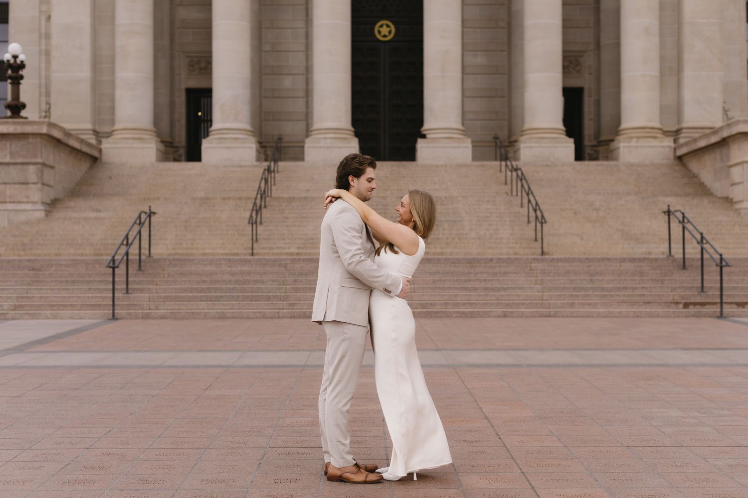 A couple in light-colored formal attire embraces in front of a grand building with stone columns and steps.