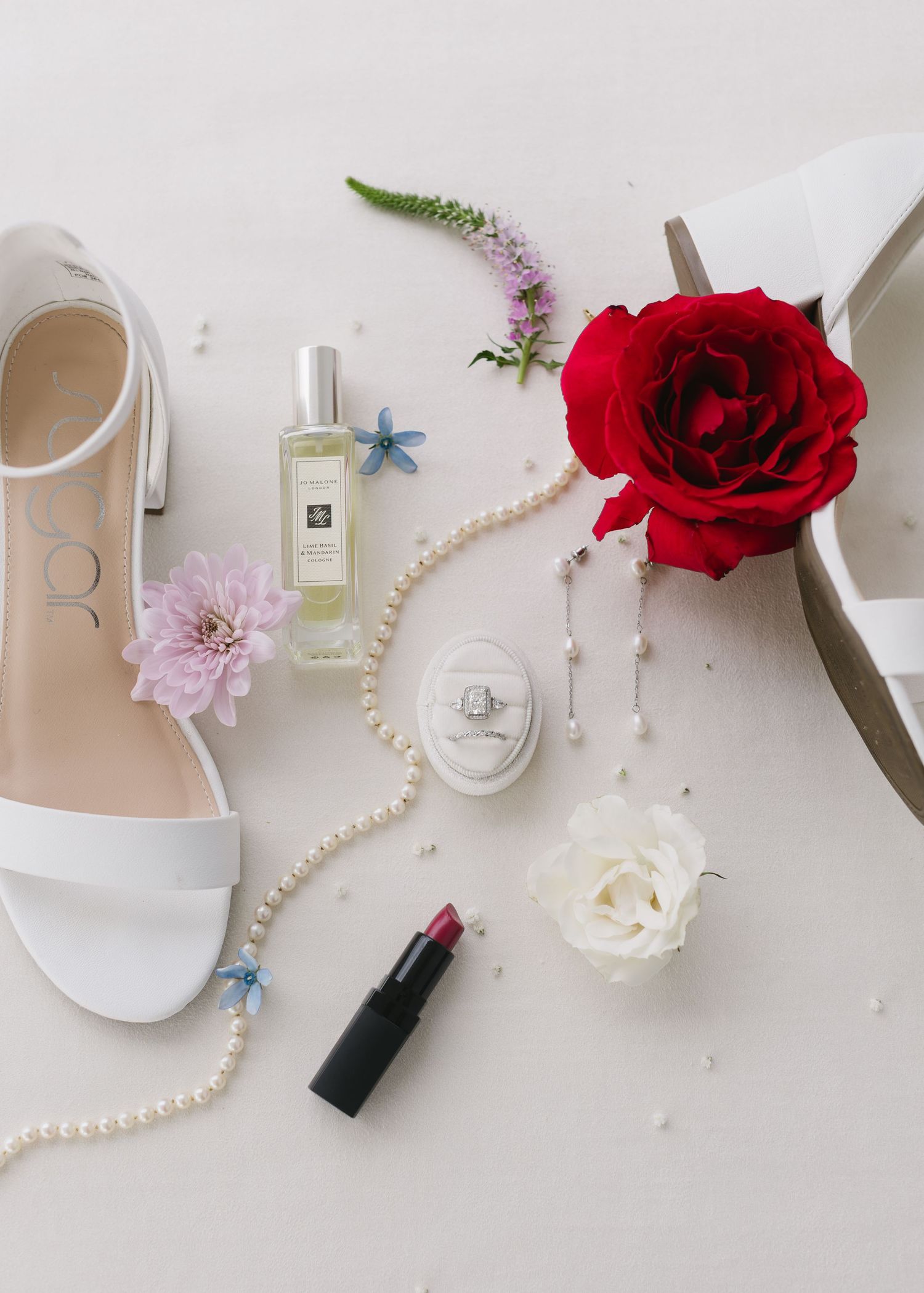 Flatlay of wedding accessories including white sandals, red roses, pearls, perfume bottles and delicate flowers on white surface.