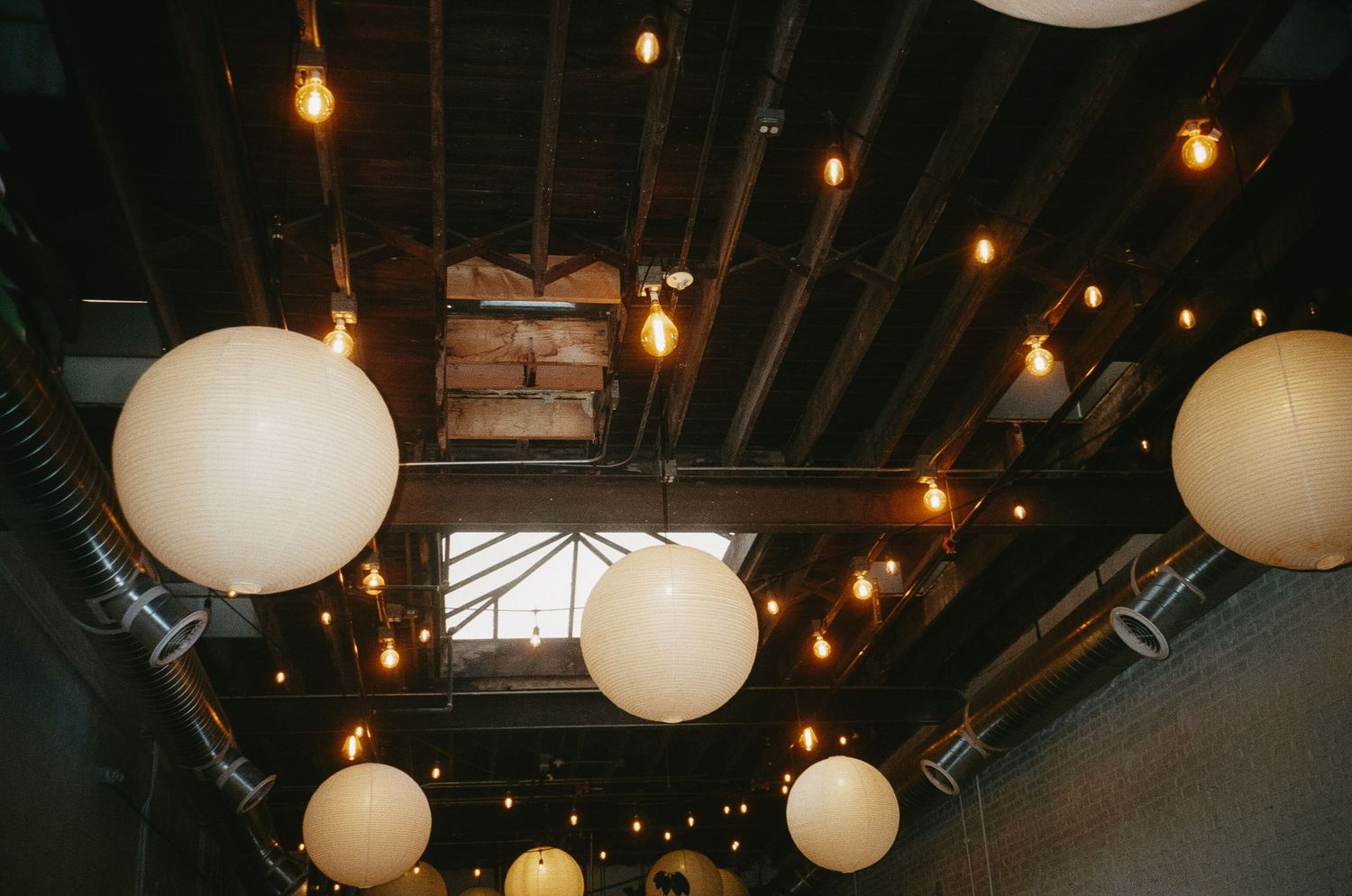 Industrial-style ceiling with exposed beams features hanging string lights and white paper lanterns creating ambient lighting.