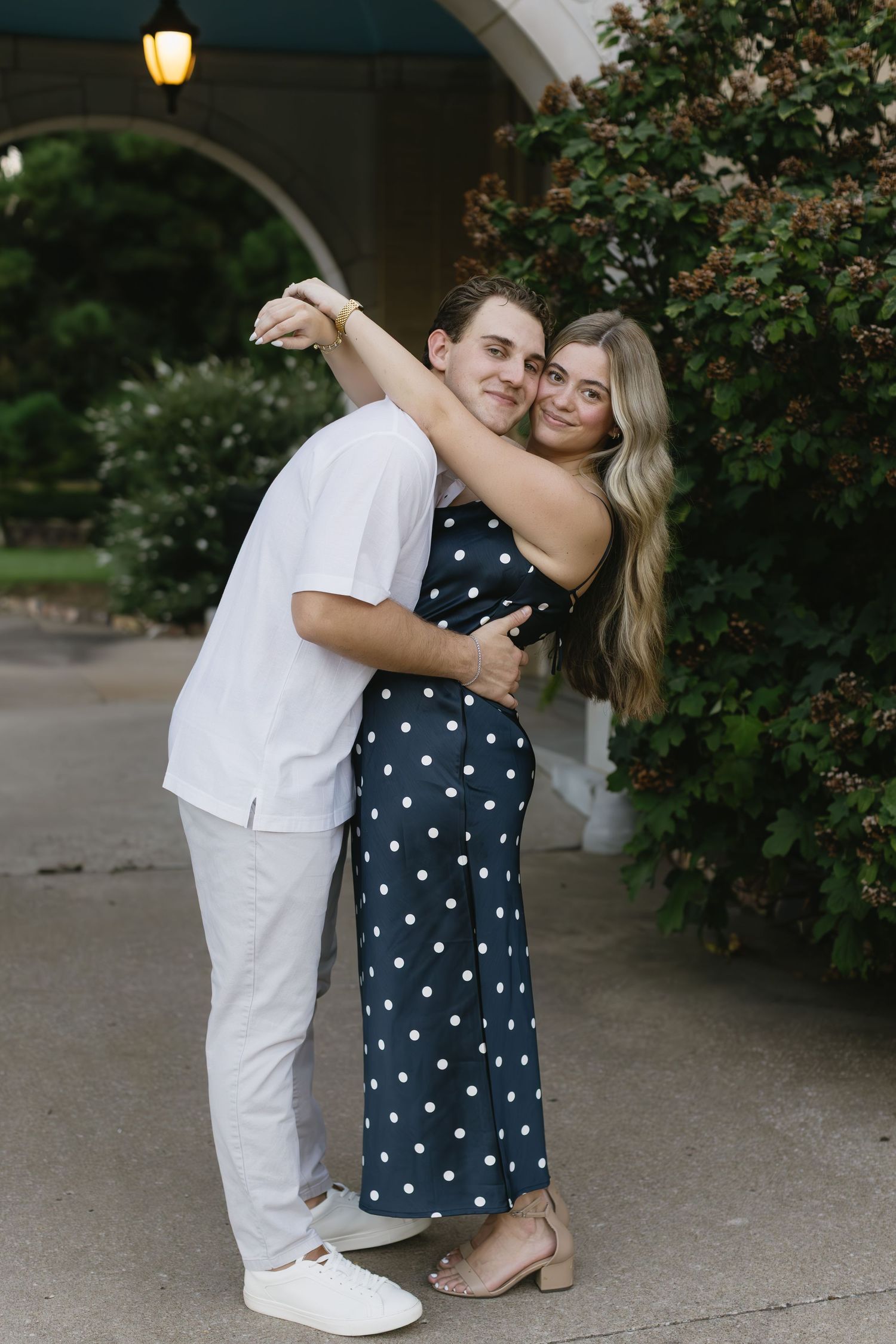 A couple in navy polka dot top and white shirt share playful romantic moments outdoors near garden bushes.