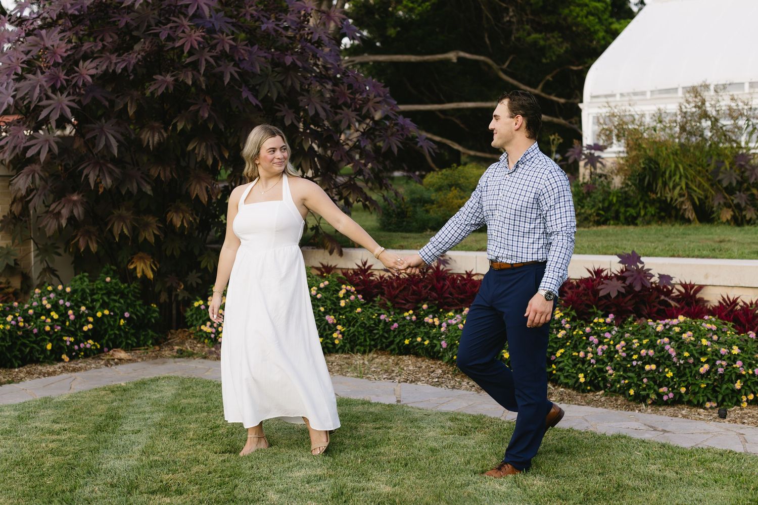Couple in white dress and checkered shirt pose playfully in blooming garden setting.