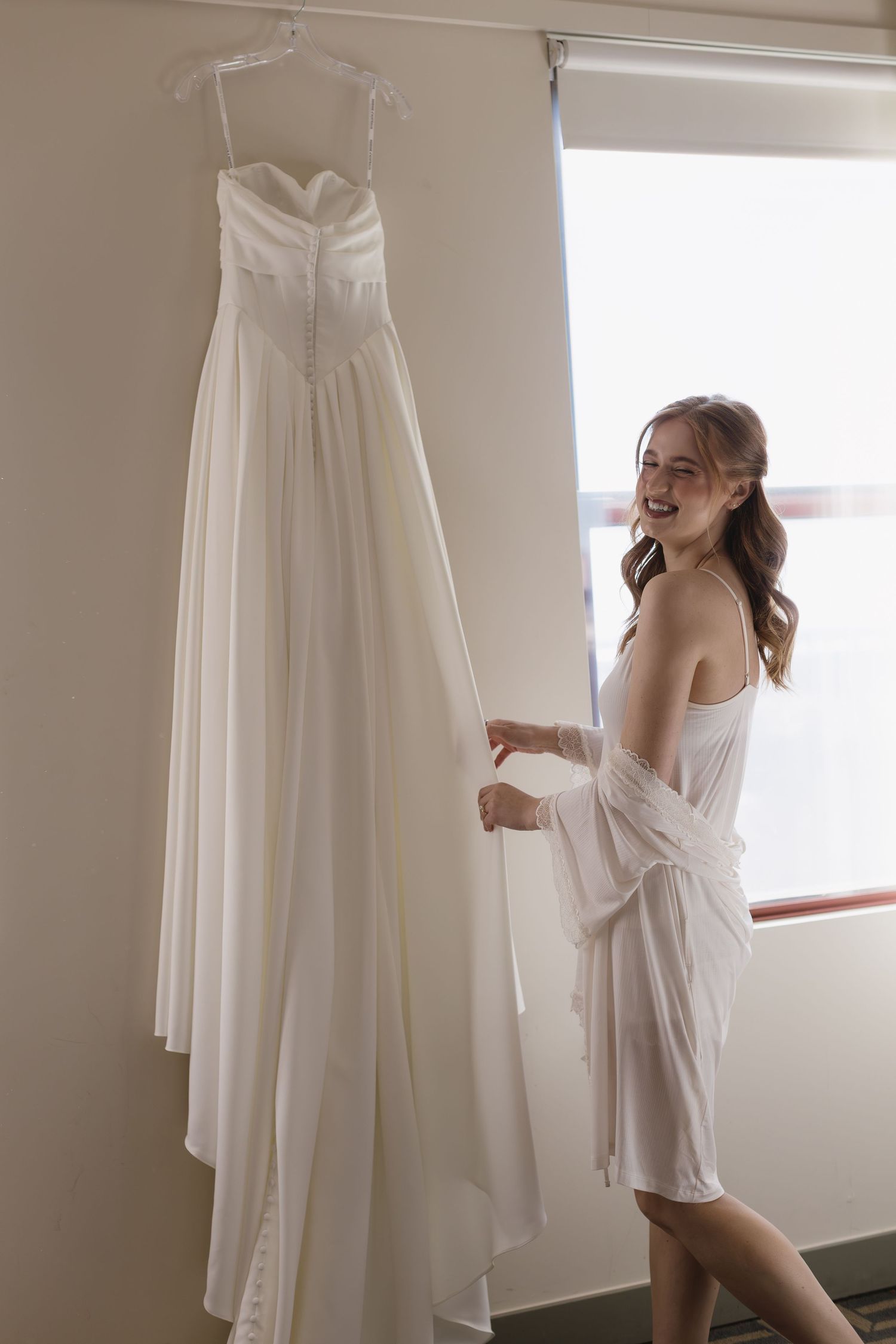 Elegant white wedding dress hanging while someone in a white robe admires it by a window.