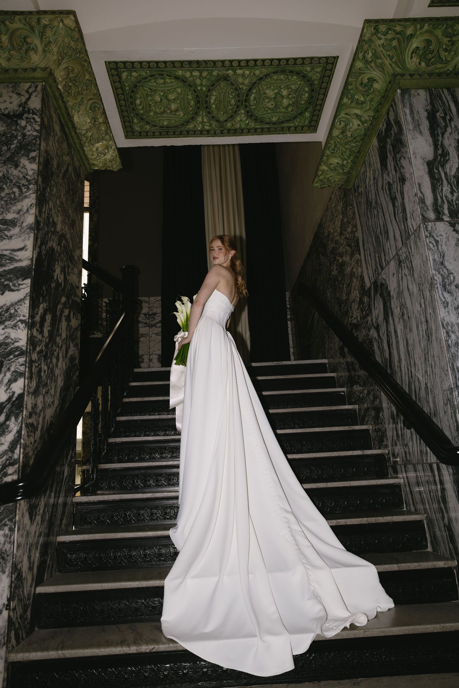 A bride in a flowing white wedding gown with a long train stands on a grand staircase with ornate green walls.