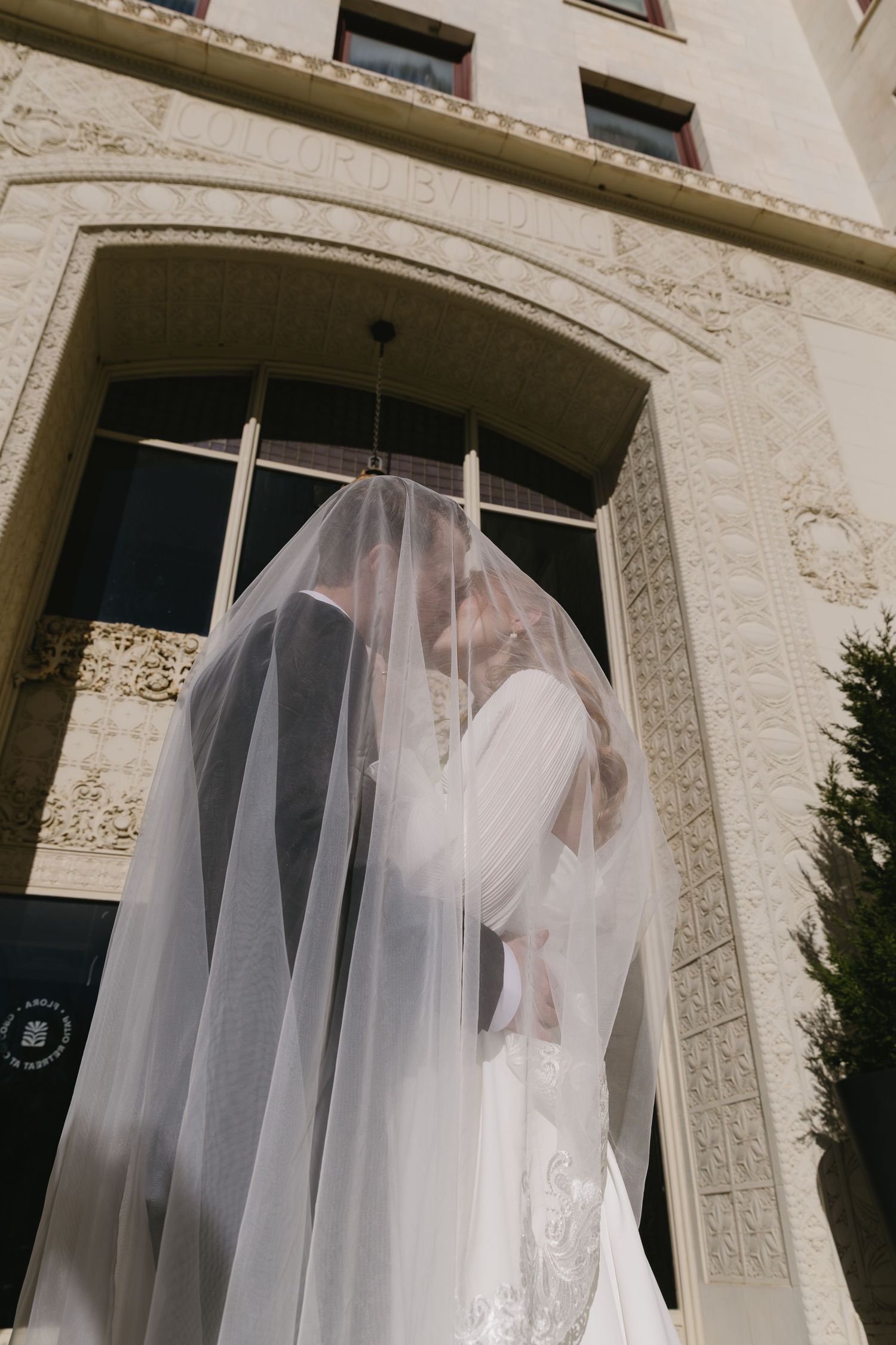 Wedding veil flowing in the breeze against stone building exterior.