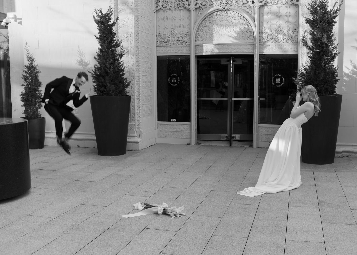 Playful black and white wedding photo with couple and fallen bouquet outside venue entrance.