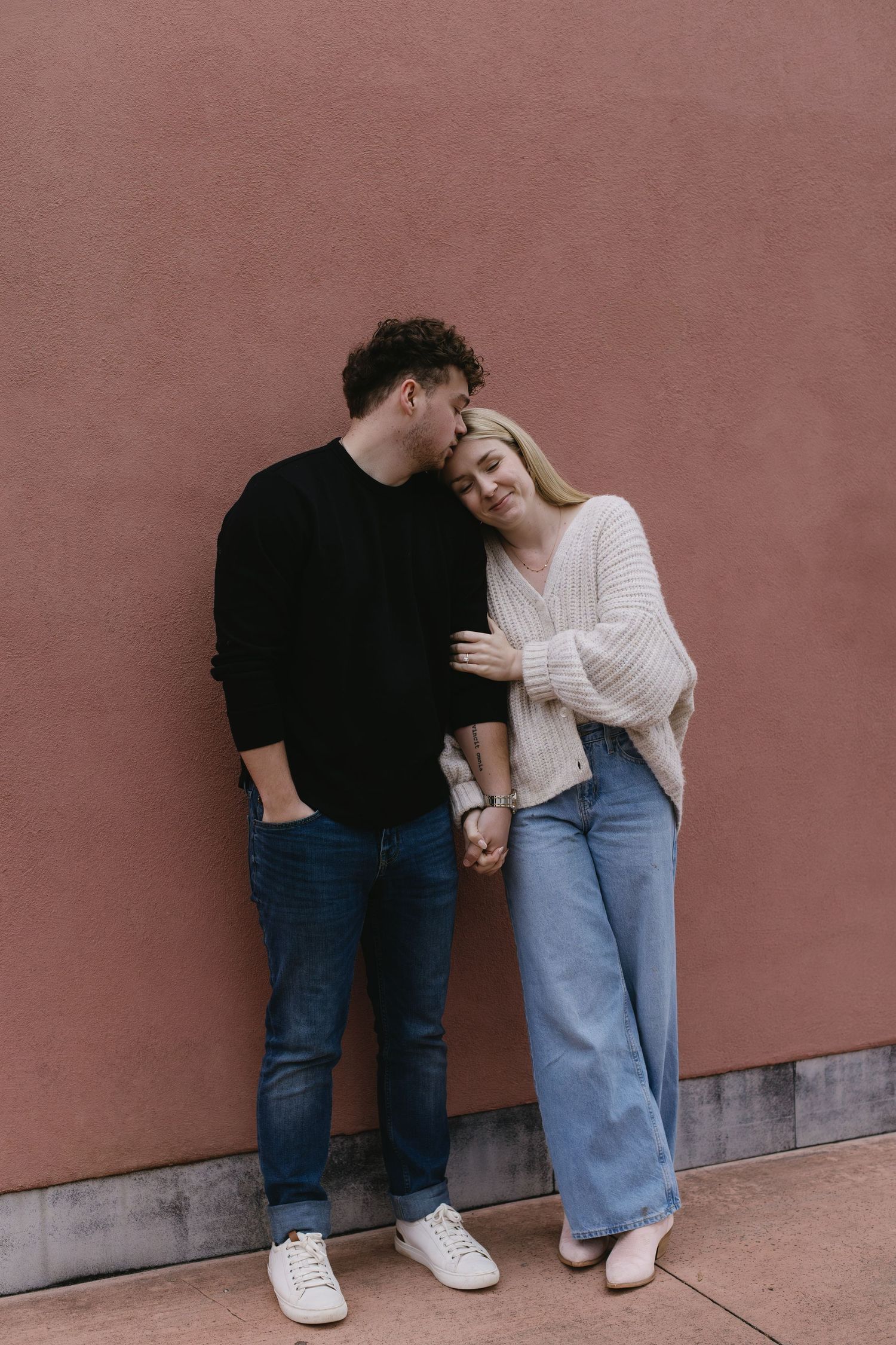 A couple in jeans and casual wear posing playfully against a pink wall.