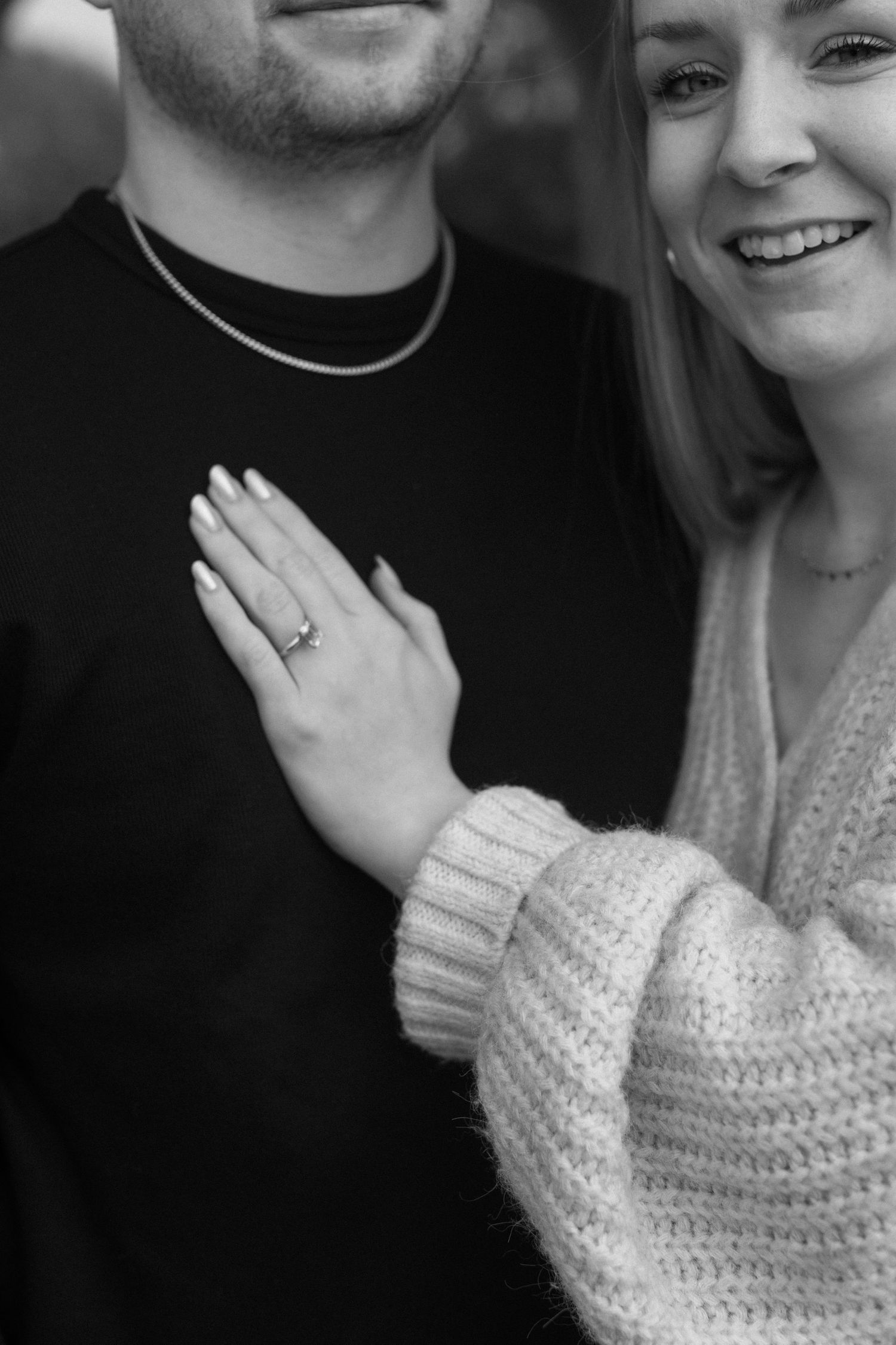 A close-up black and white engagement photo showing a diamond ring on a hand against a cozy sweater.