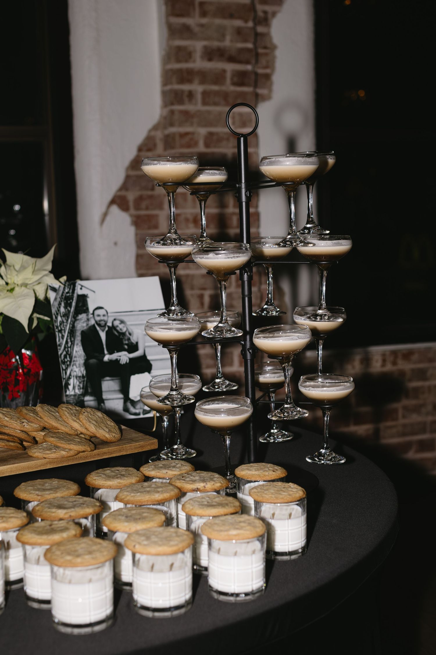 Wedding dessert display featuring tiered champagne glasses and cookies on black tablecloth against brick wall.
