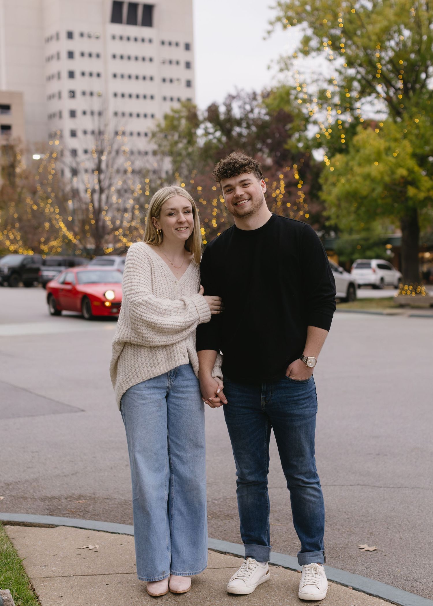 A couple in casual wear standing together on a city sidewalk with buildings in background.