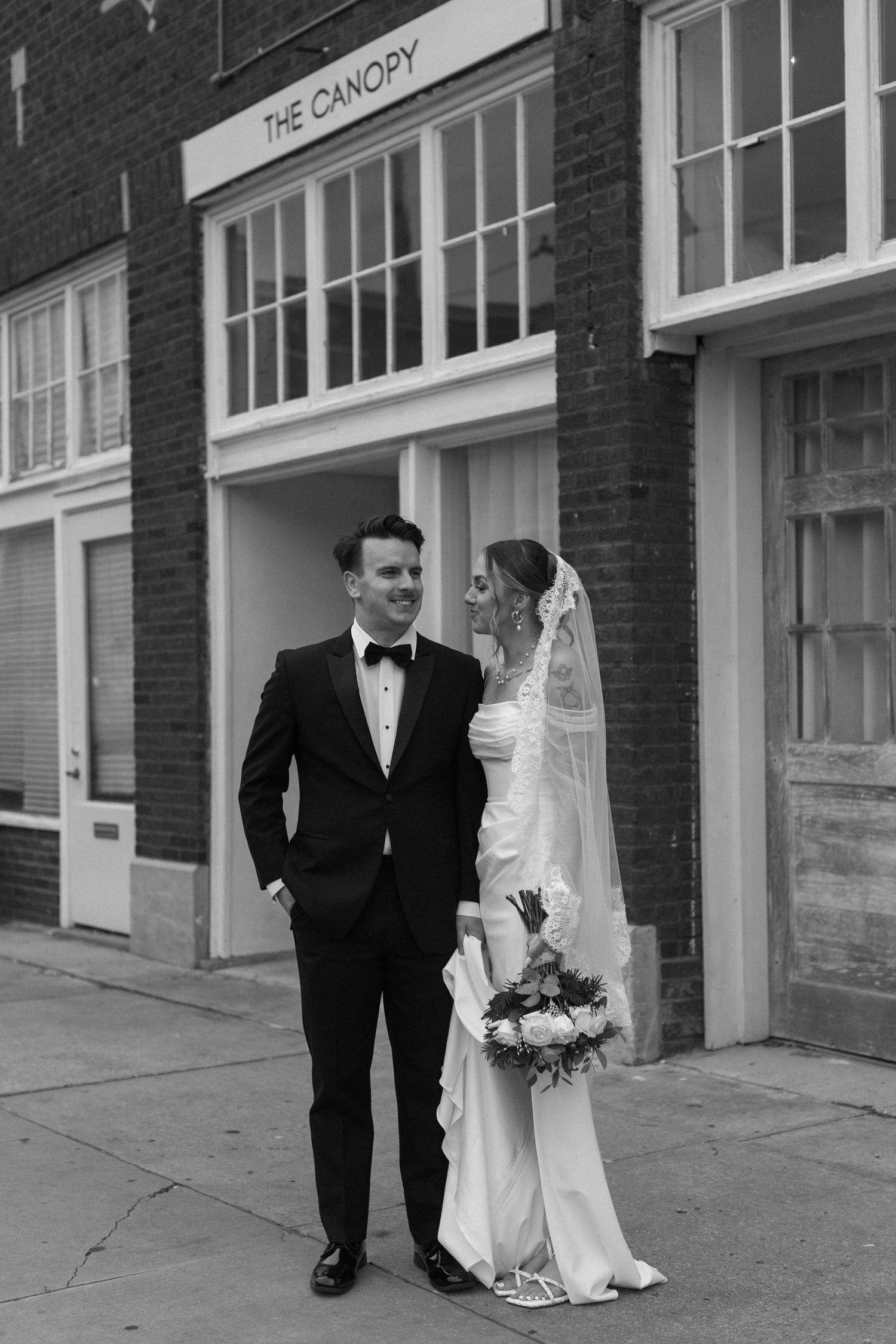 Black and white wedding photo of couple standing outside brick storefront on urban sidewalk.
