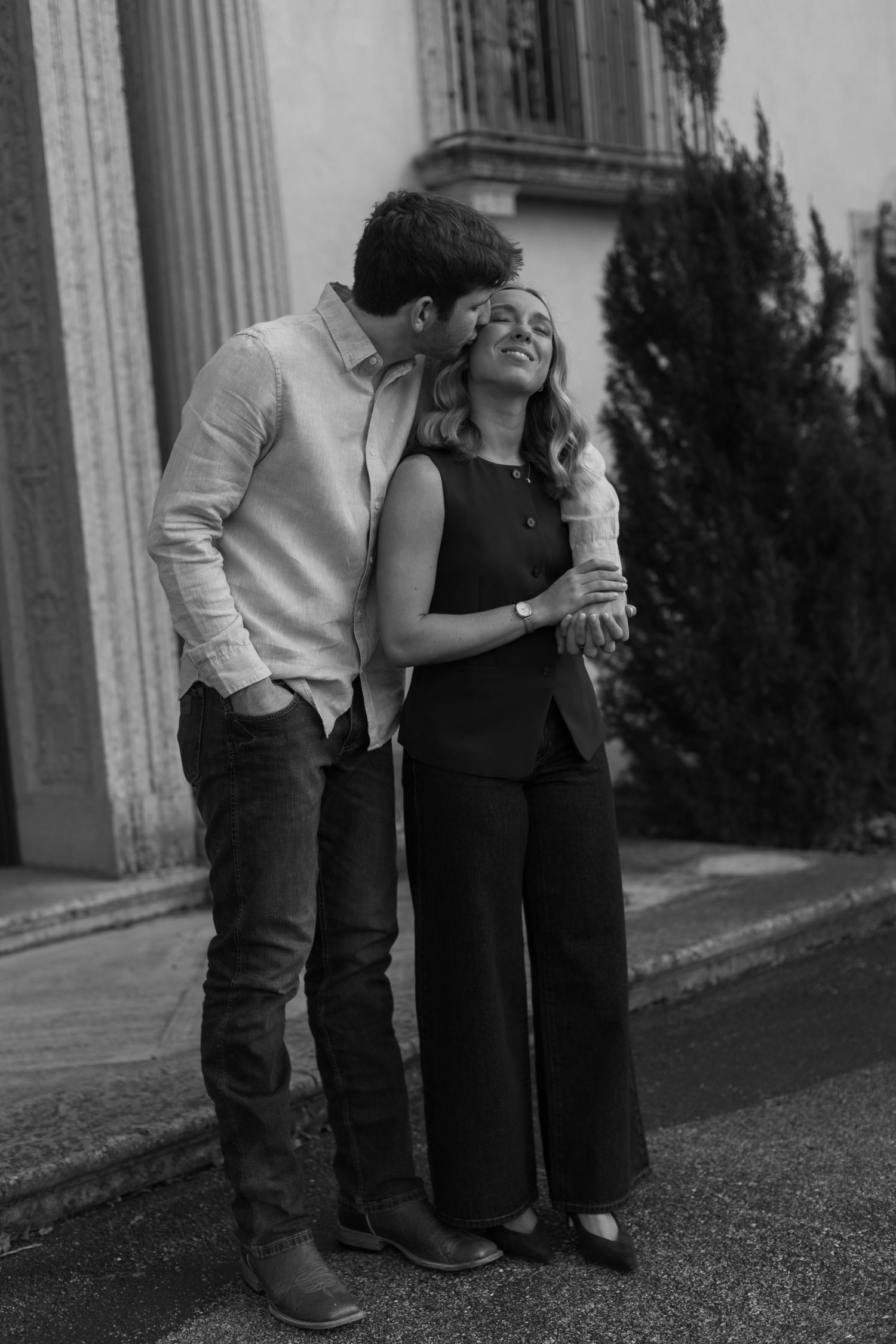 A couple laughs together while standing against a classical building exterior.