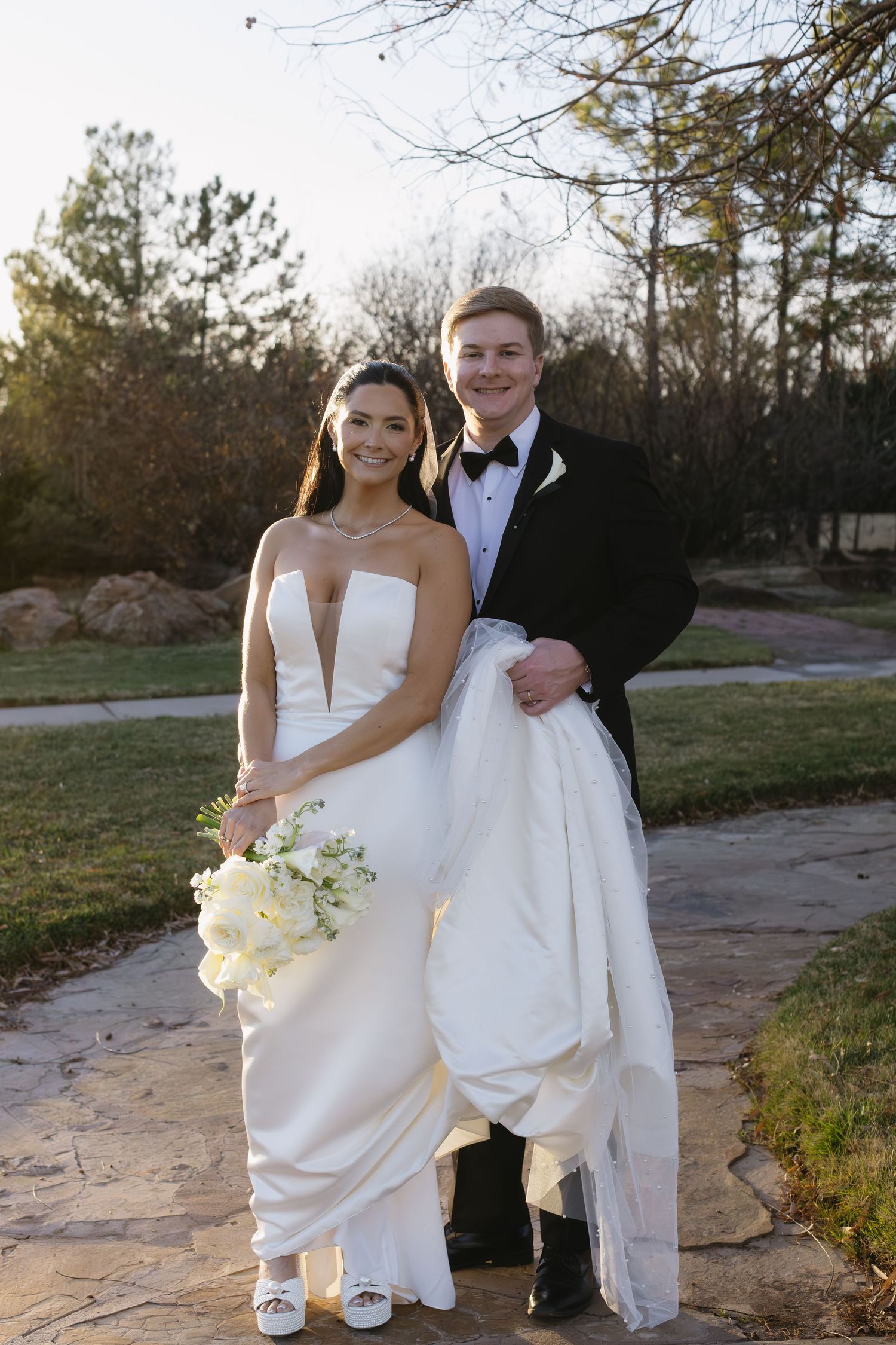Wedding couple in formal attire pose outdoors with white floral bouquet at sunset.