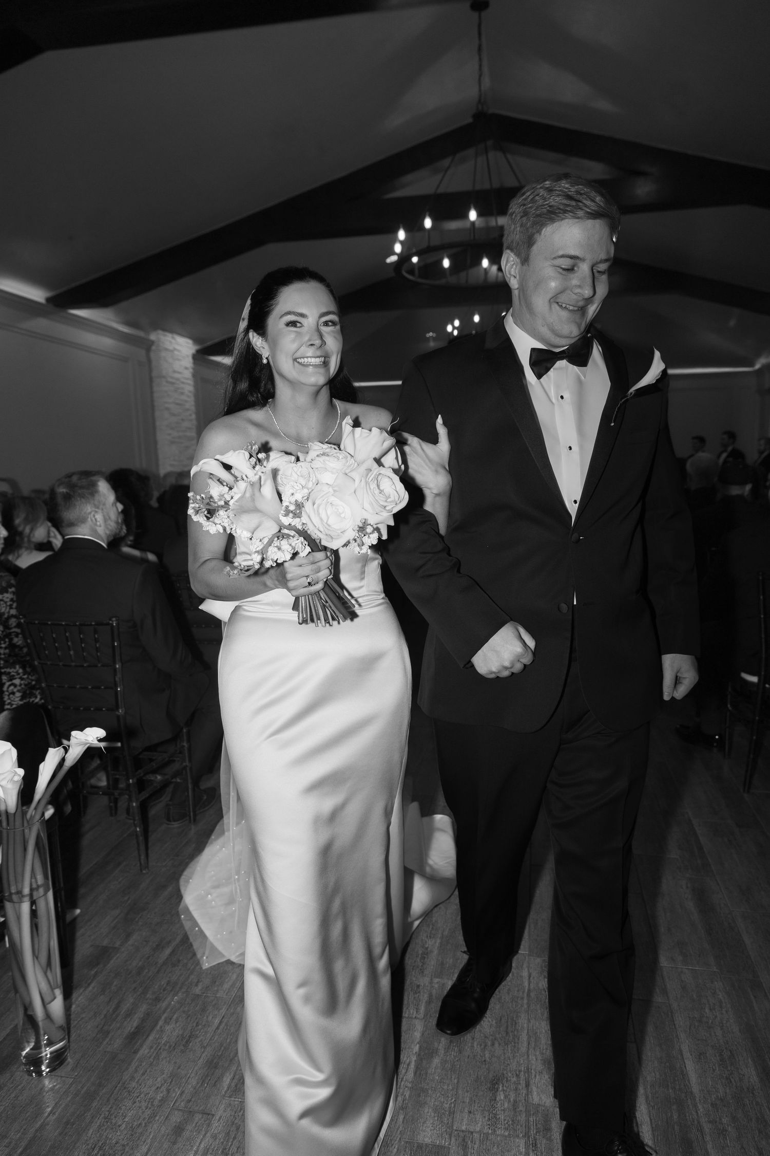 A couple walks down the aisle at their wedding, with the bride in white holding a bouquet and groom in classic tuxedo.
