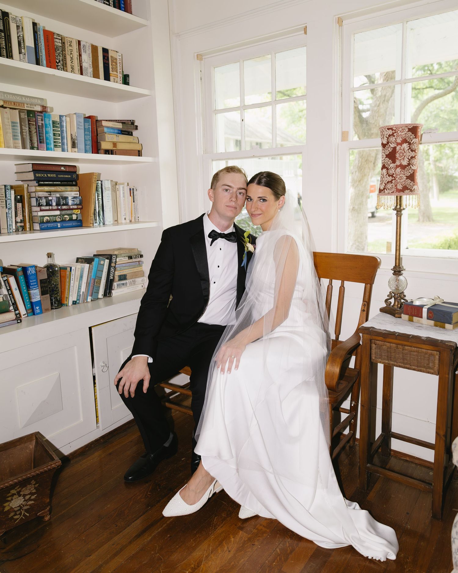A romantic wedding photo series showing a couple in a library with white bookshelves and wooden chairs.