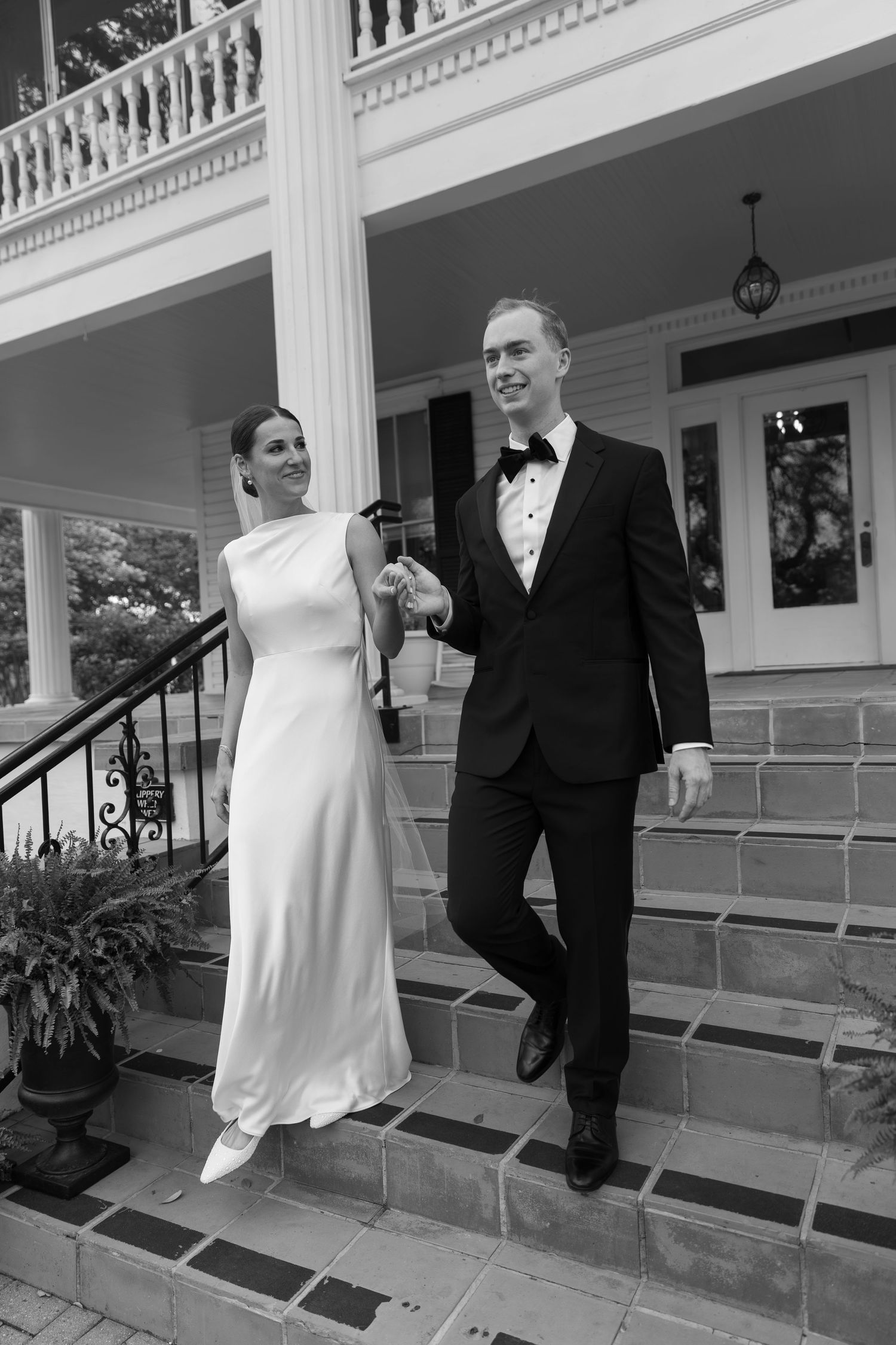 A happy couple shares candid moments on the steps of a grand white-pillared porch in formal wedding attire.
