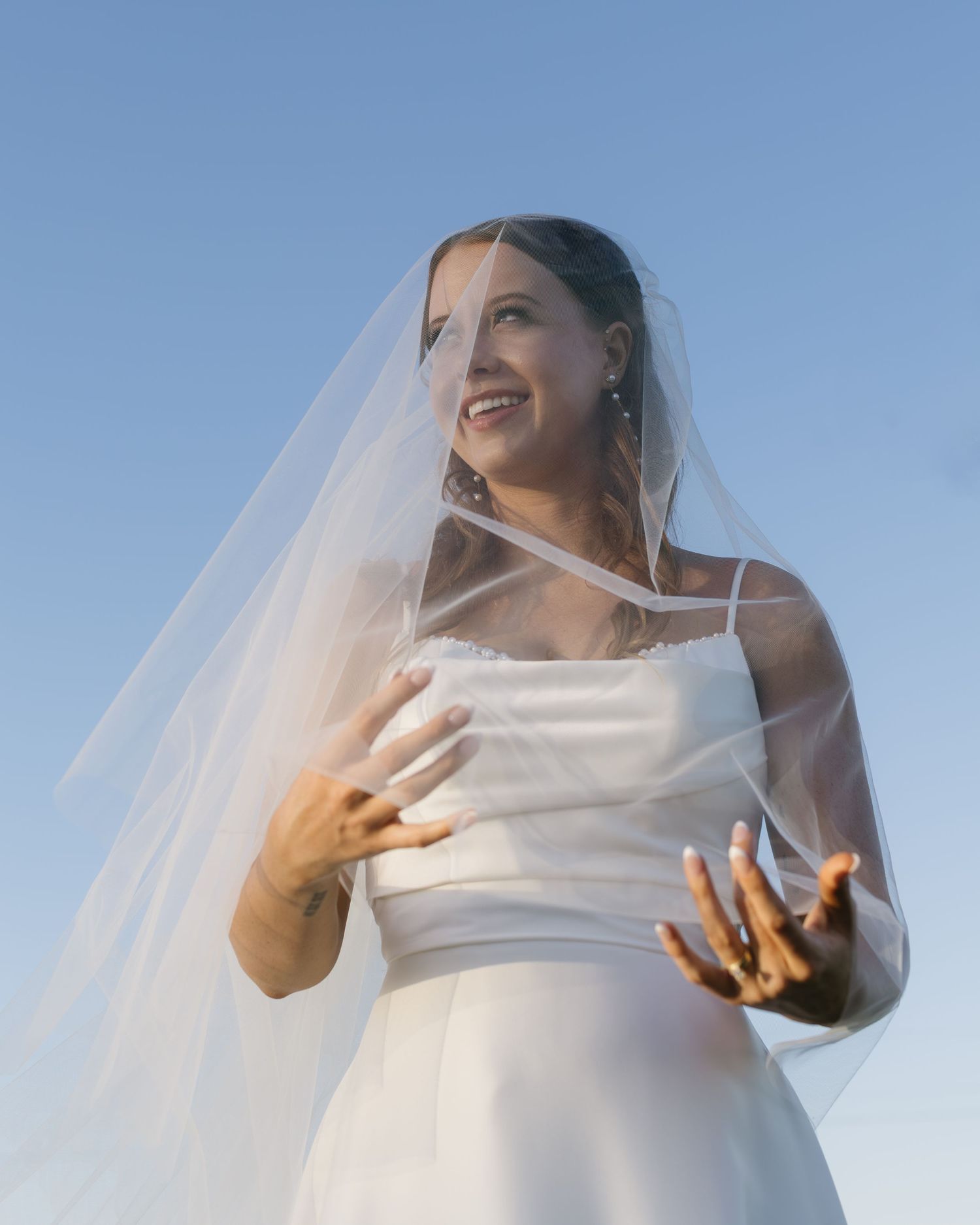 A bride in a sleek white dress gestures expressively against a bright blue sky.