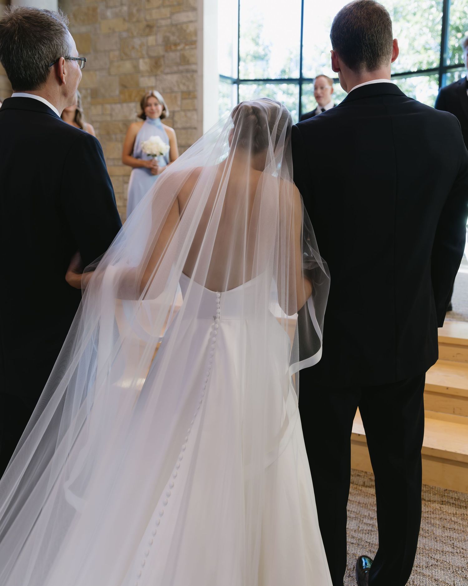 Bride in flowing white gown walks with wedding party in formal procession.