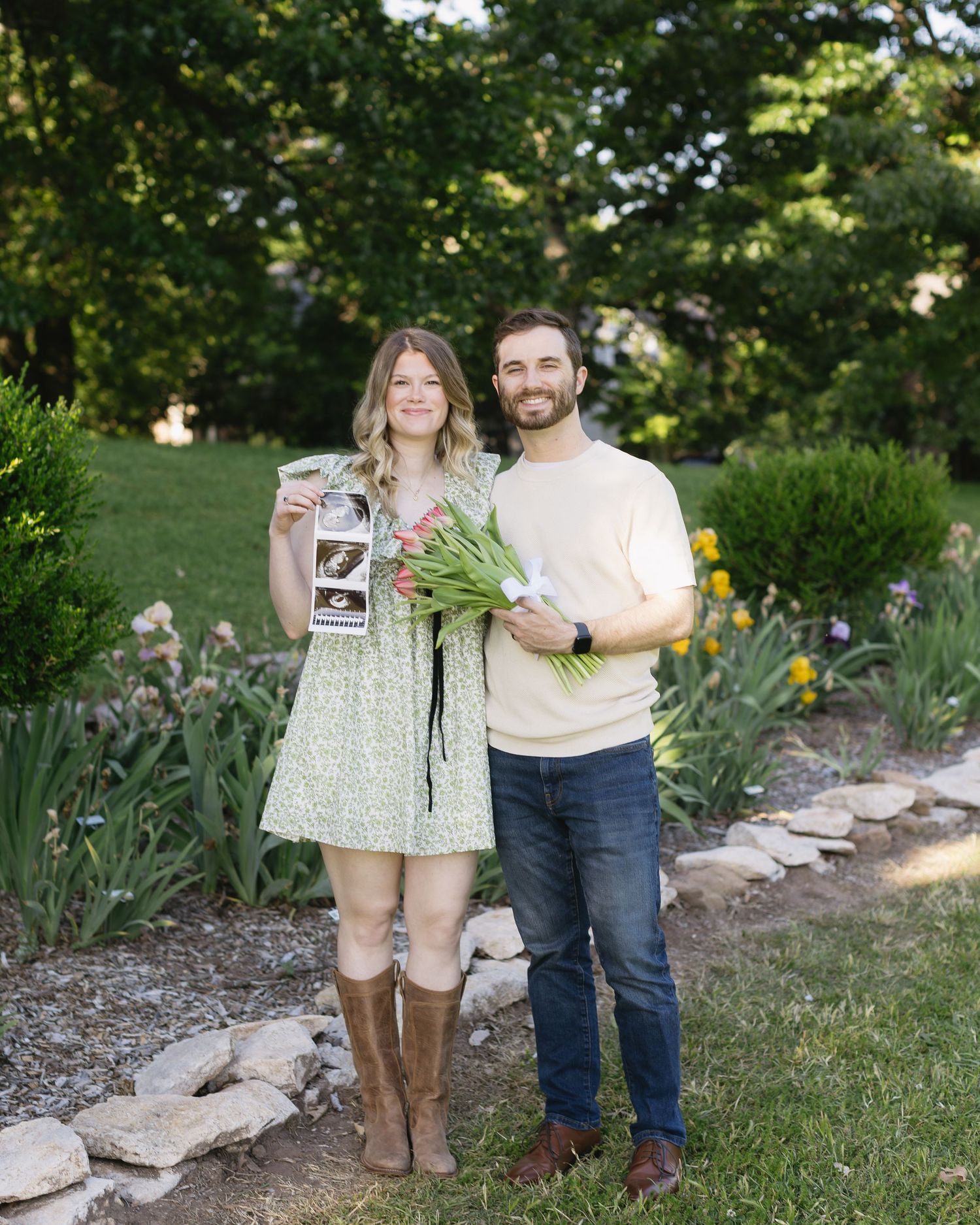 Couple pose with sonogram photos and flowers in a garden setting with stone pathway.
