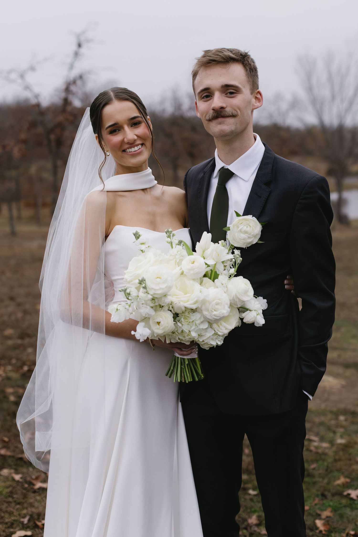 Newlyweds pose outdoors with white floral bouquet against autumn landscape.
