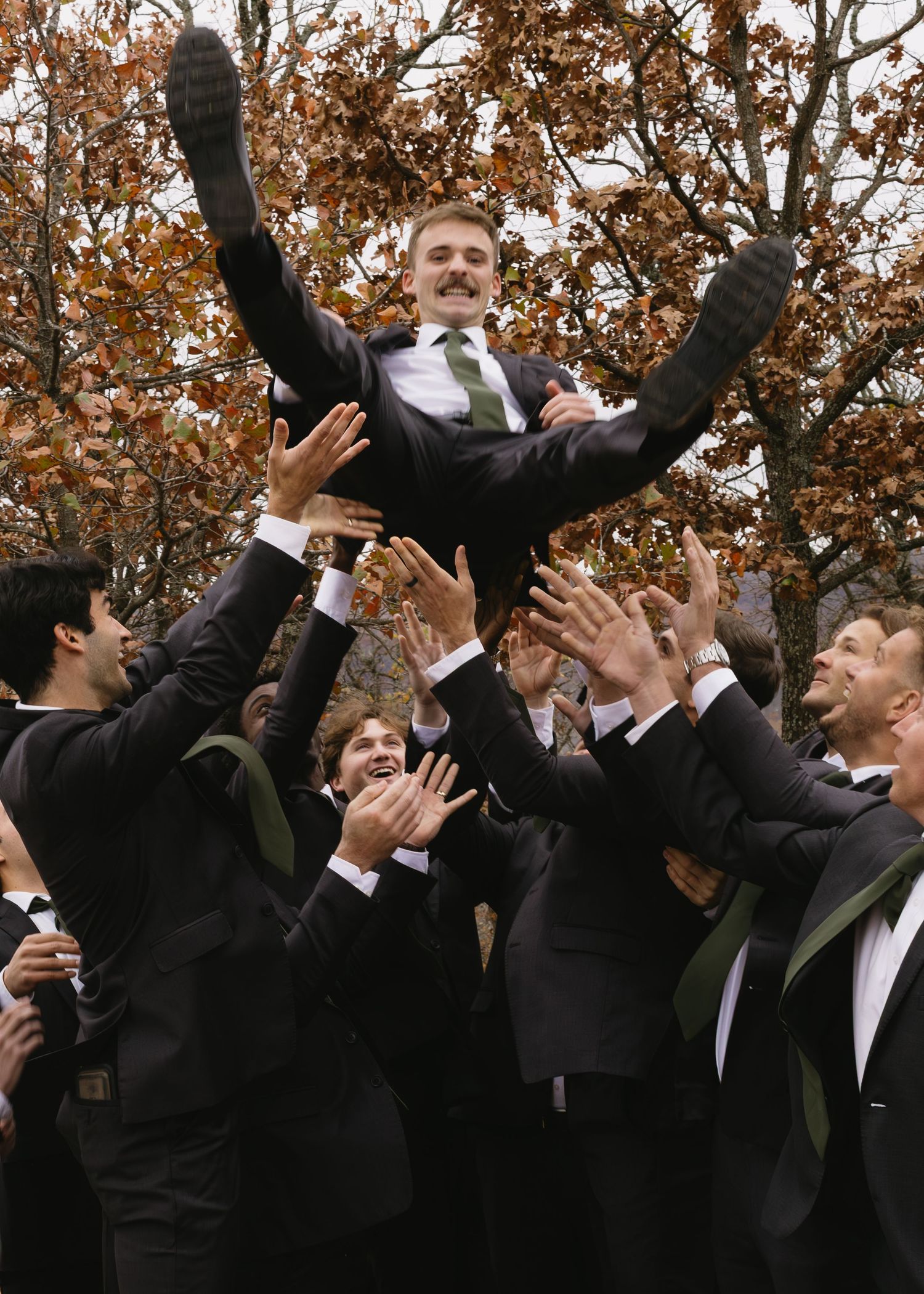 Groomsmen in black suits jumping with excitement during outdoor autumn wedding celebration.