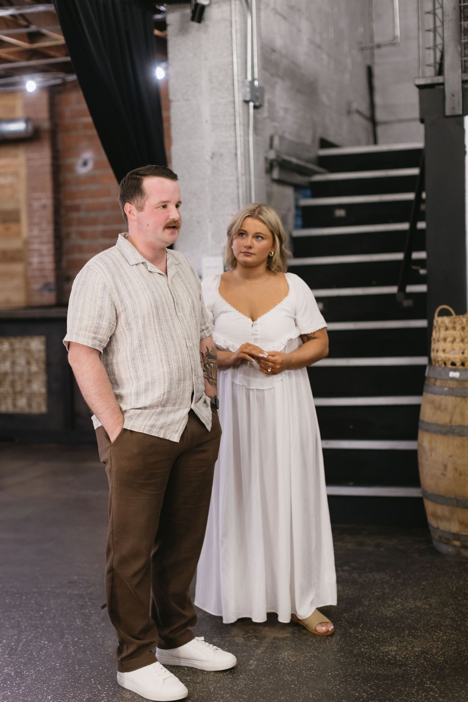 Couple standing together in industrial-style venue with wine barrels and staircase.