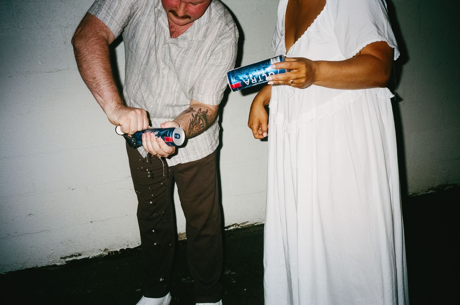 People in wedding attire examining something in dim lighting against white wall.