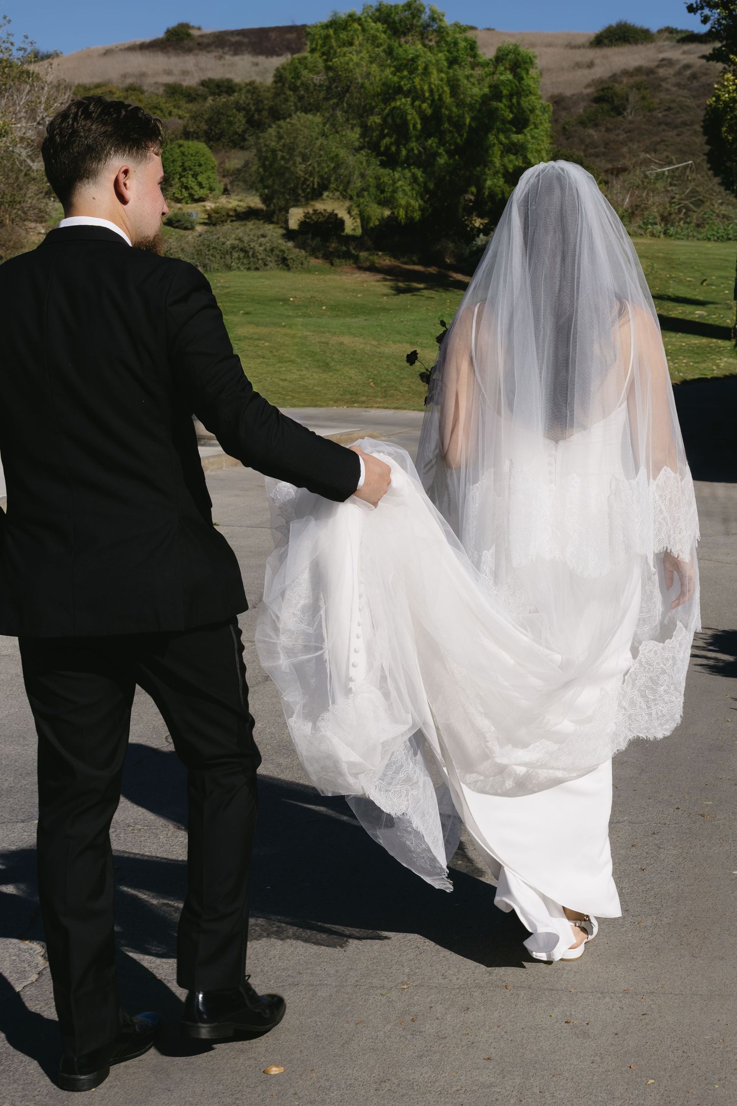 Wedding couple walking together on a path with flowing white veil trailing behind them.